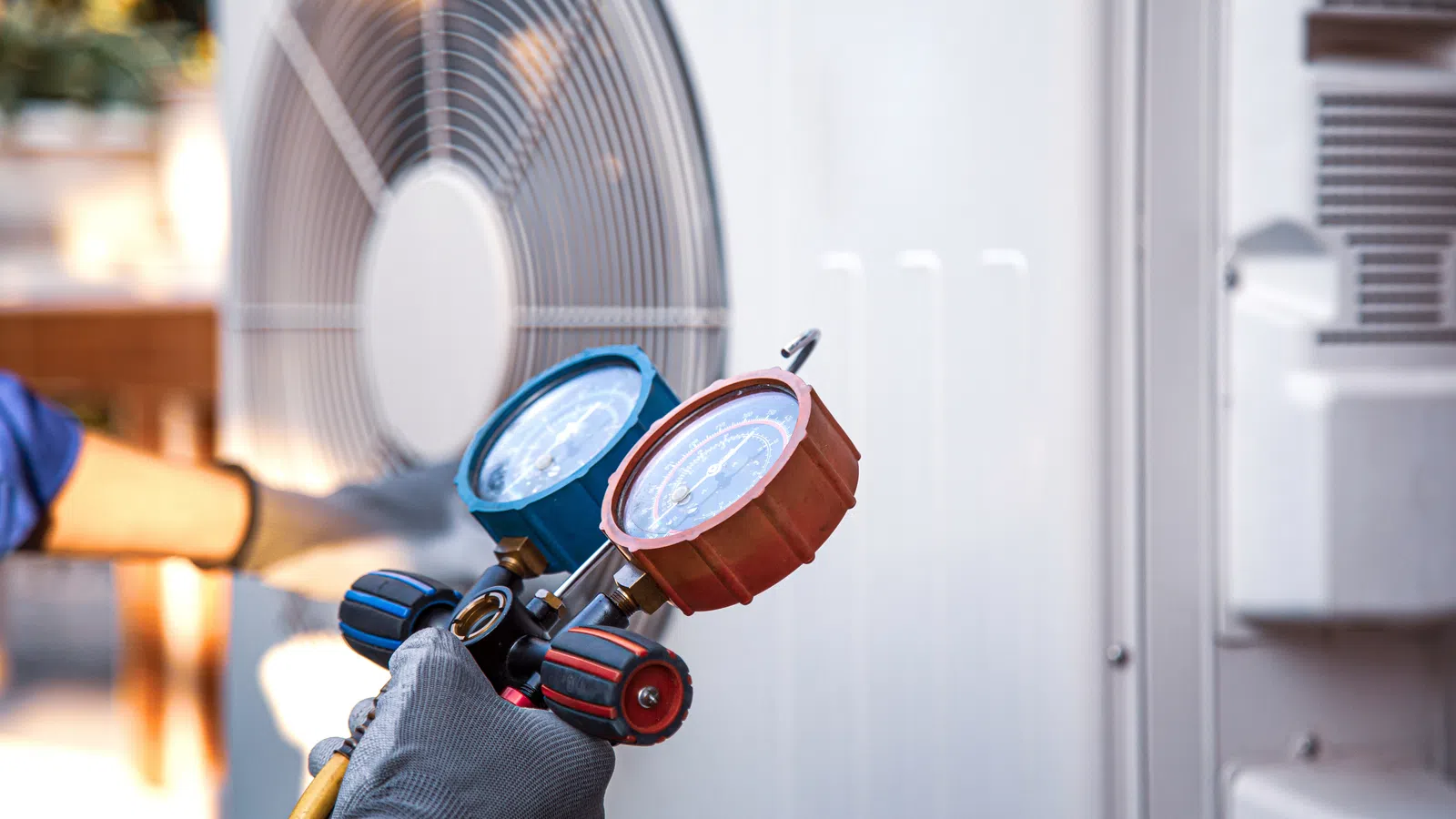 Technician's hand in glove holding HVAC manifold gauge set near outdoor air conditioning unit fan.