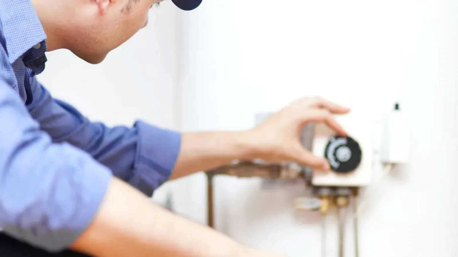 Technician adjusting controls on a white water heater in a residential setting wearing a blue shirt and cap.