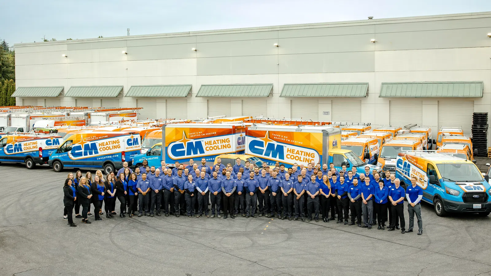 Large group of CM Heating Cooling employees posing in front of company service vans outside warehouse.