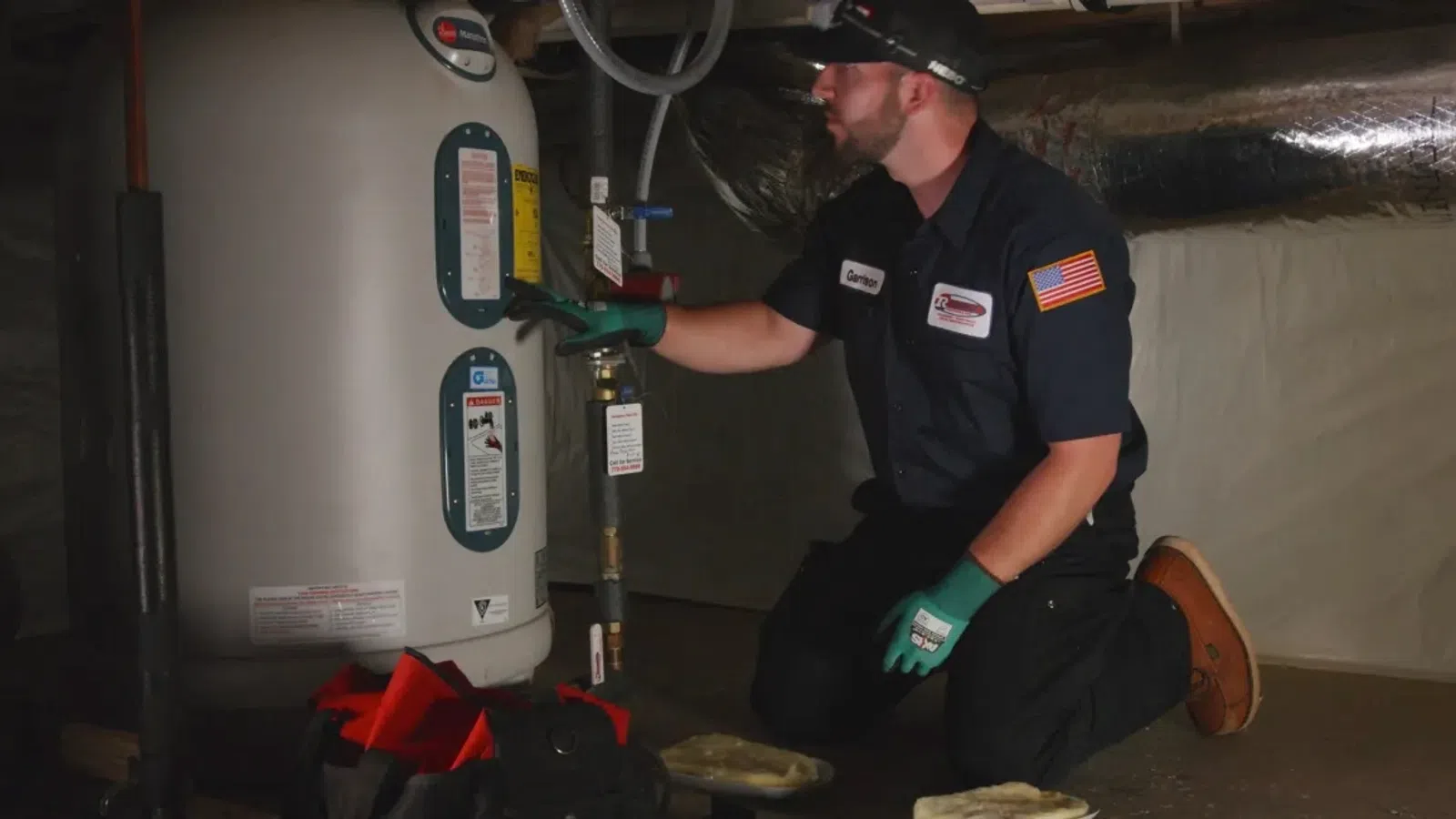 Technician kneeling by a water heater, inspecting pipes and valves in a dimly lit basement.