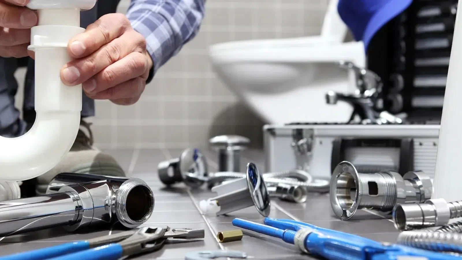 Plumber assembling white PVC pipe with metal plumbing tools and bathroom fixtures around a toilet in the background