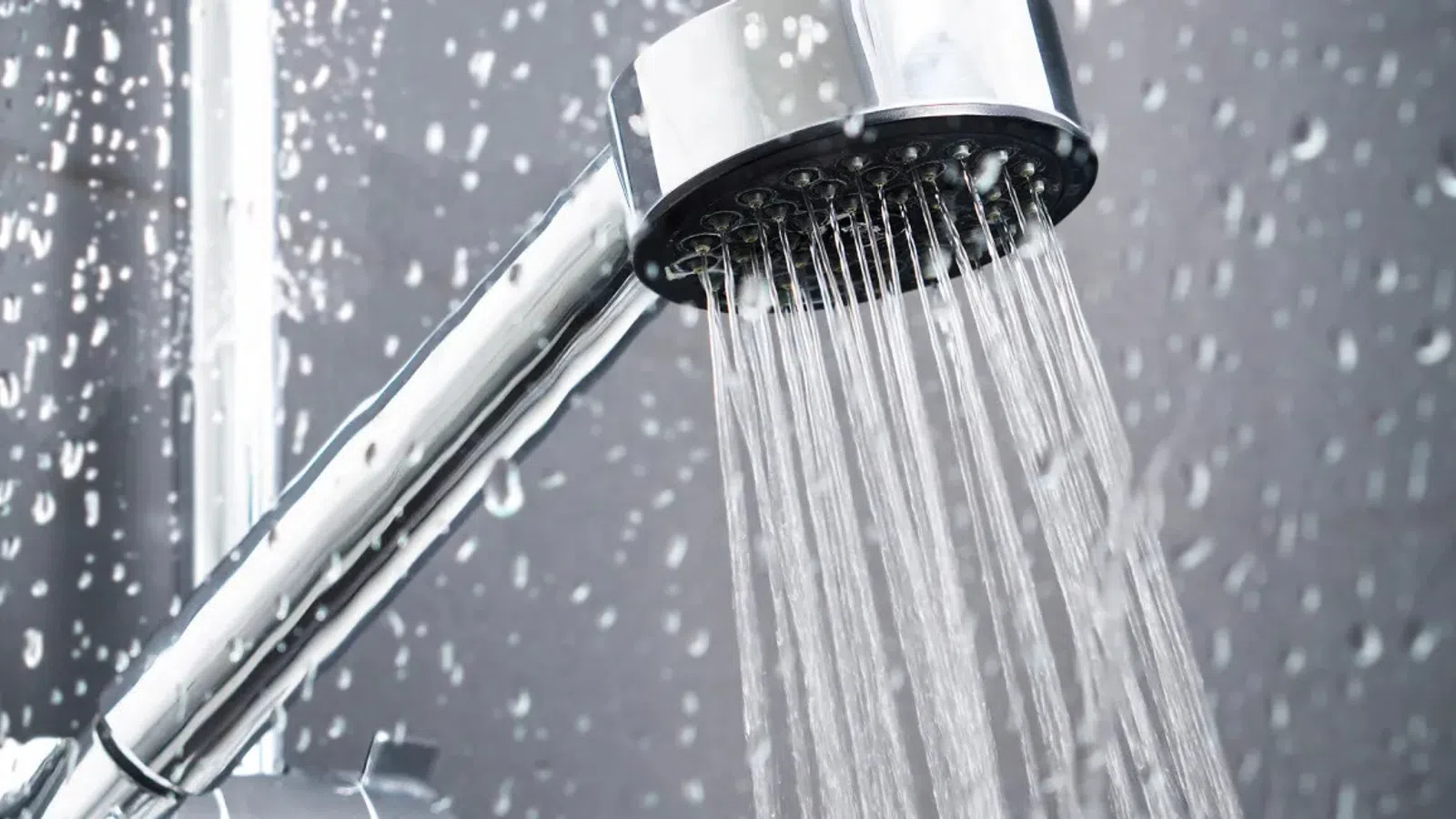 Close-up of a modern chrome showerhead releasing a steady stream of water droplets against a gray tiled background