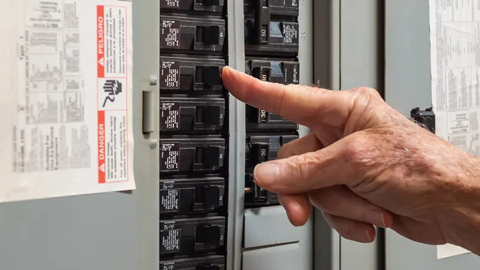 Hand adjusting circuit breaker switches inside an electrical panel with safety labels and technical diagrams.