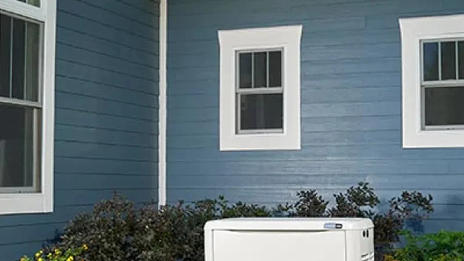 White outdoor generator installed beside blue house with white trim and garden plants around it.