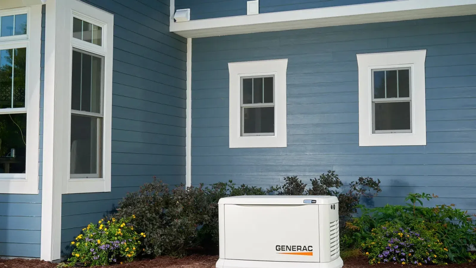 Generac home standby generator installed beside blue house with white trim and landscaped garden bed.