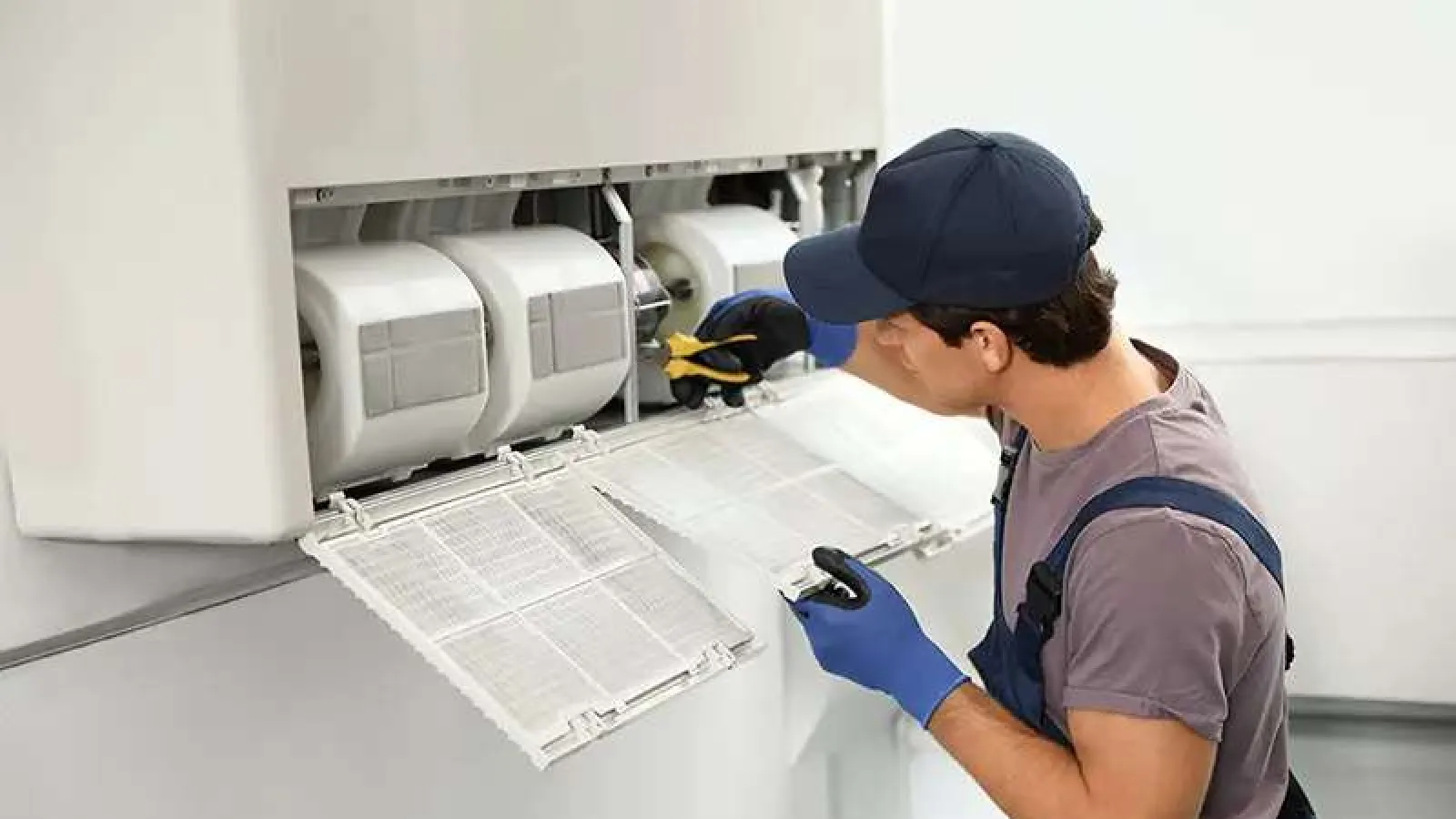 Technician wearing gloves and cap fixing HVAC air conditioning unit with tools indoors.