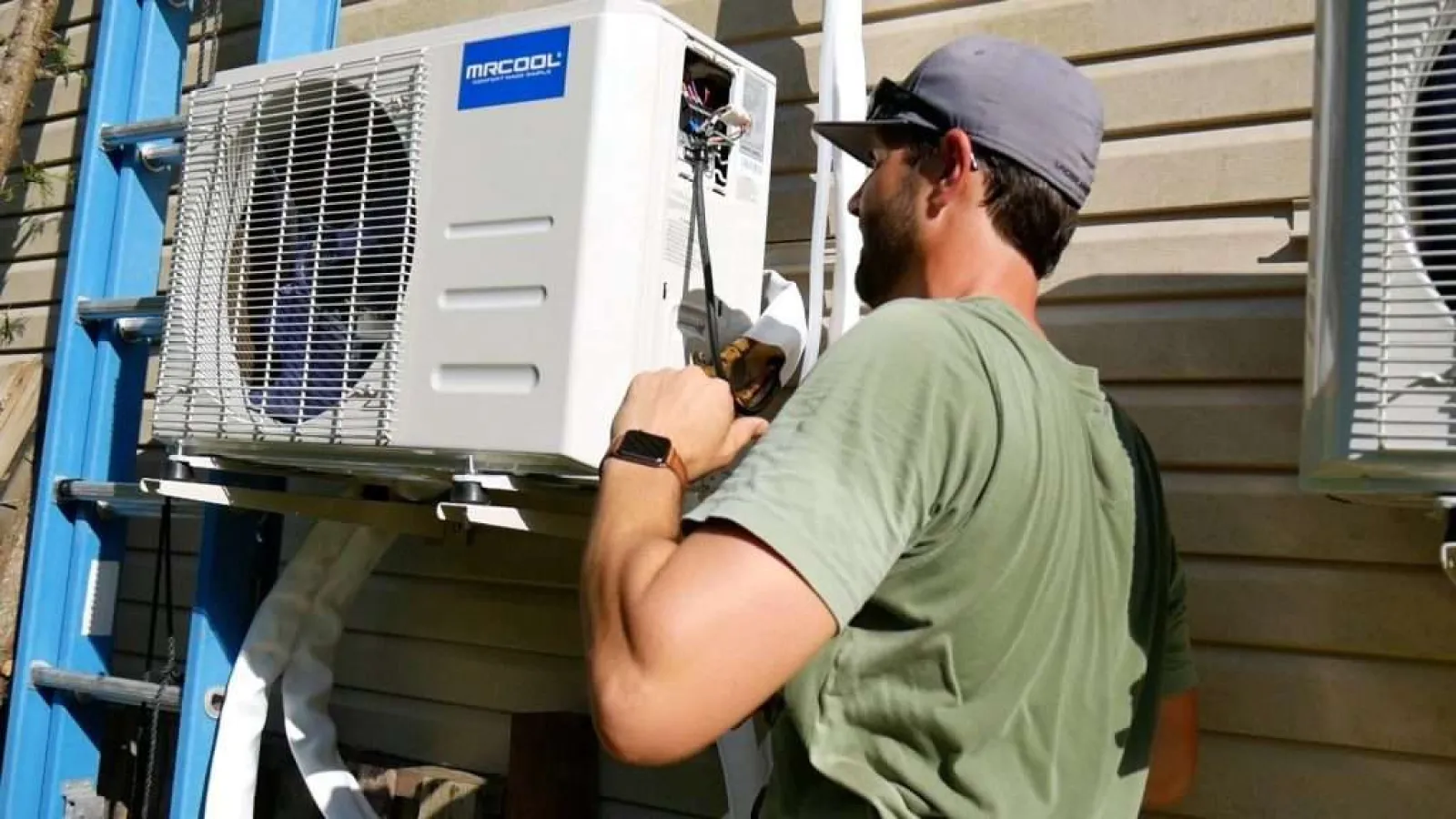 Technician in green shirt installing or repairing a white MRCOOL air conditioning unit outdoors.