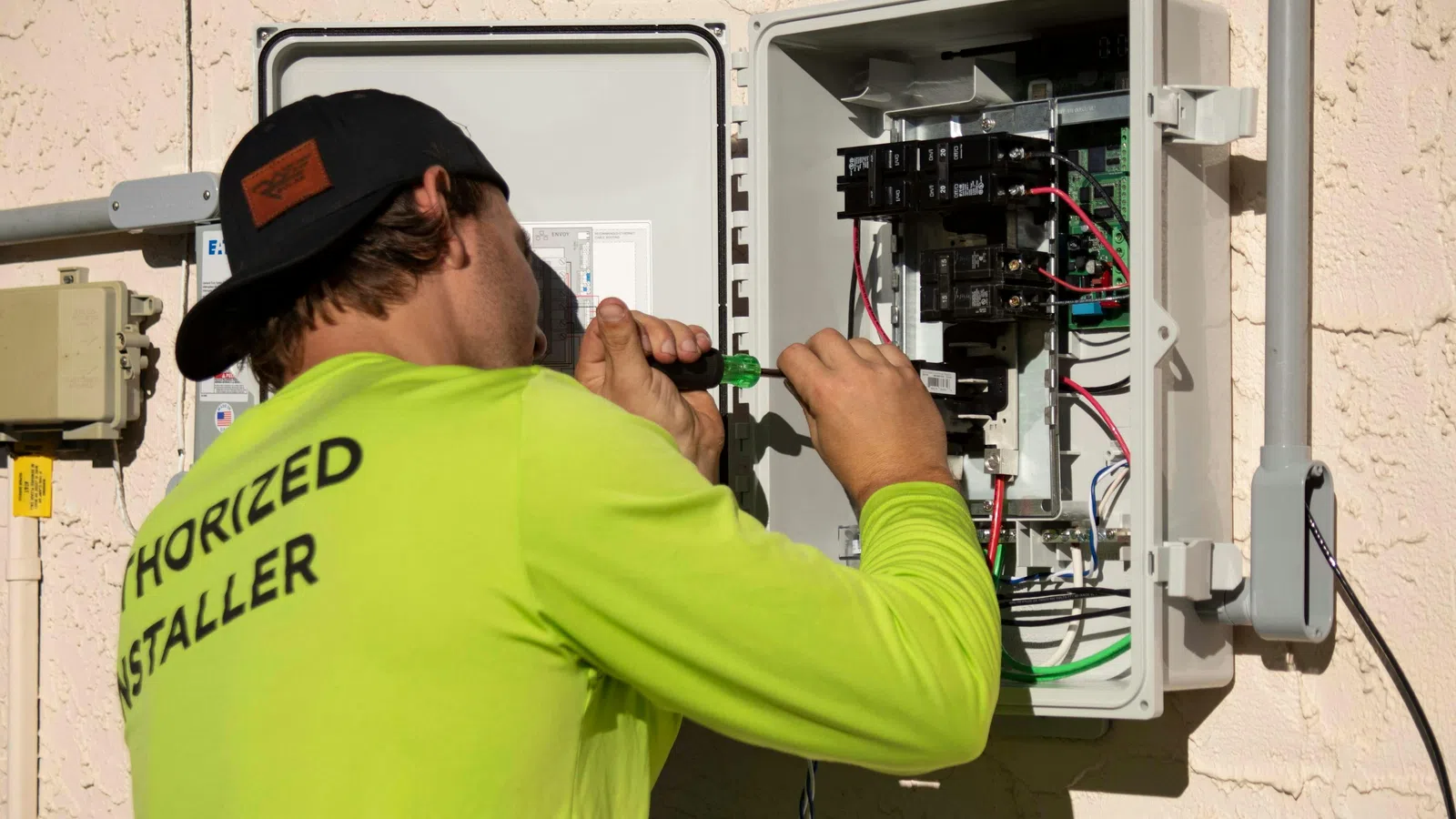 Electrician in a neon yellow shirt working on an open electrical panel with a screwdriver outdoors.