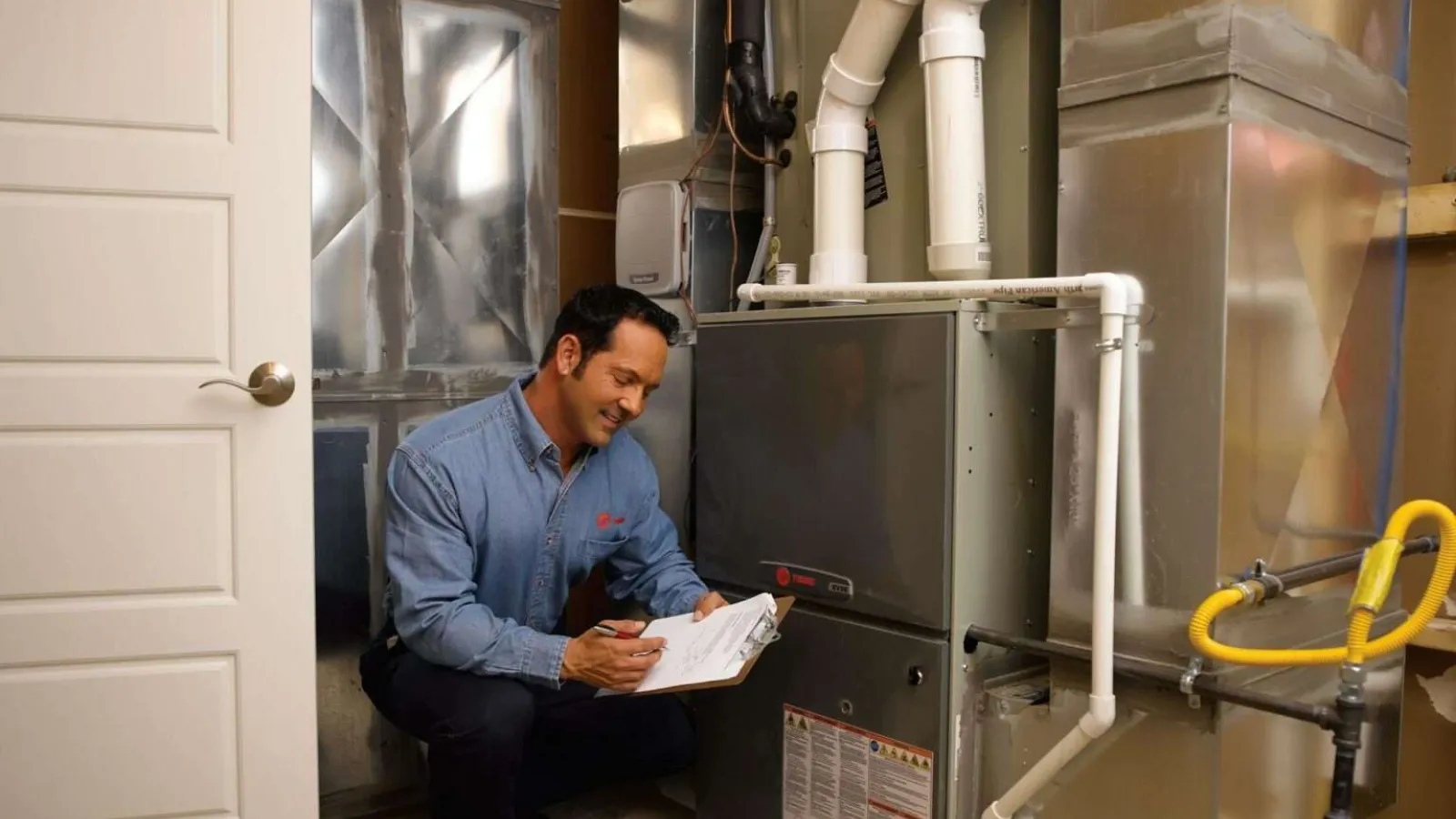 Technician inspecting and documenting details of a residential furnace in a basement with clipboard in hand