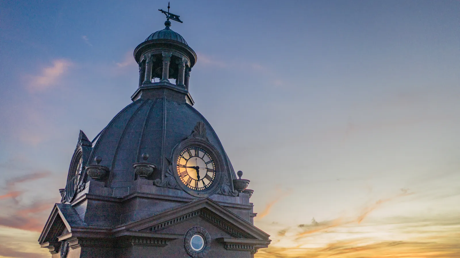 Historic clock tower with dome at sunset against a colorful sky with soft clouds and glowing horizon.
