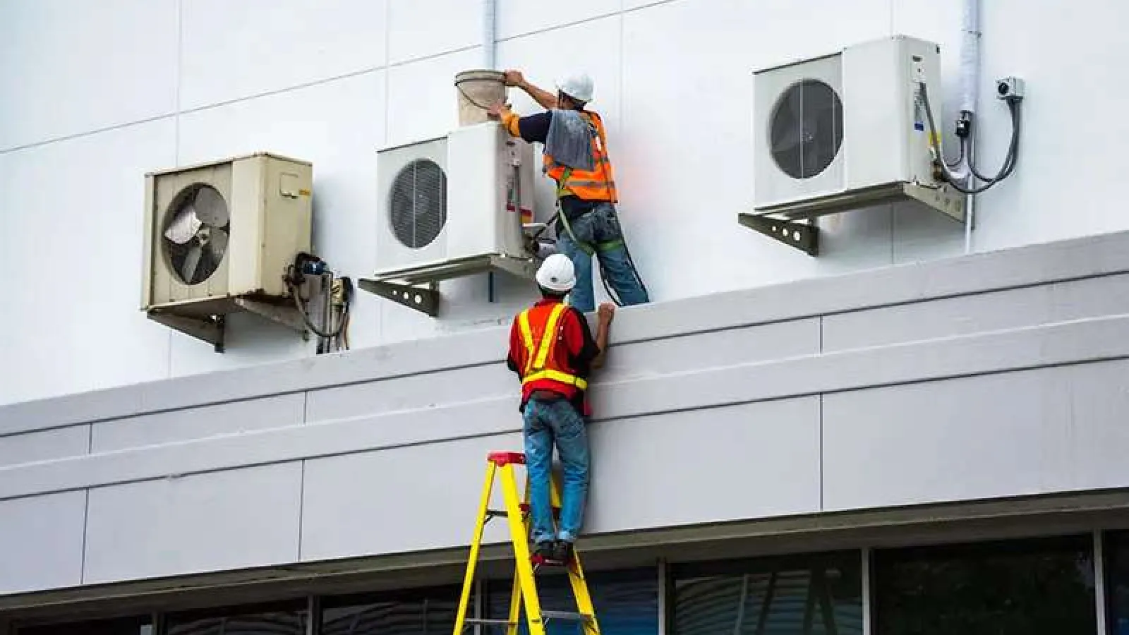 Two workers in safety gear servicing outdoor air conditioning units on a building wall using a ladder.