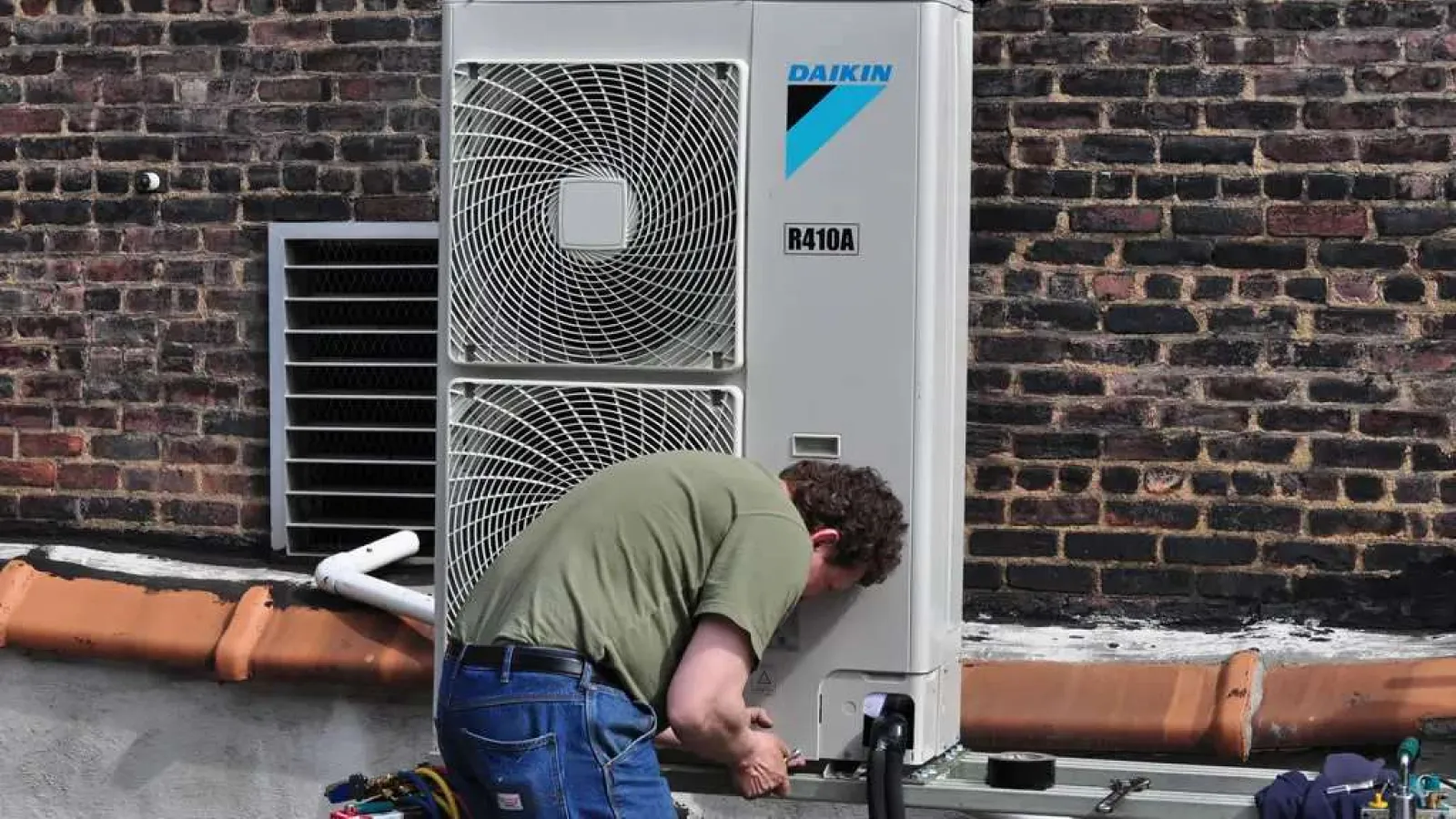 Technician repairing a Daikin R410A air conditioning unit mounted on a rooftop with brick wall background