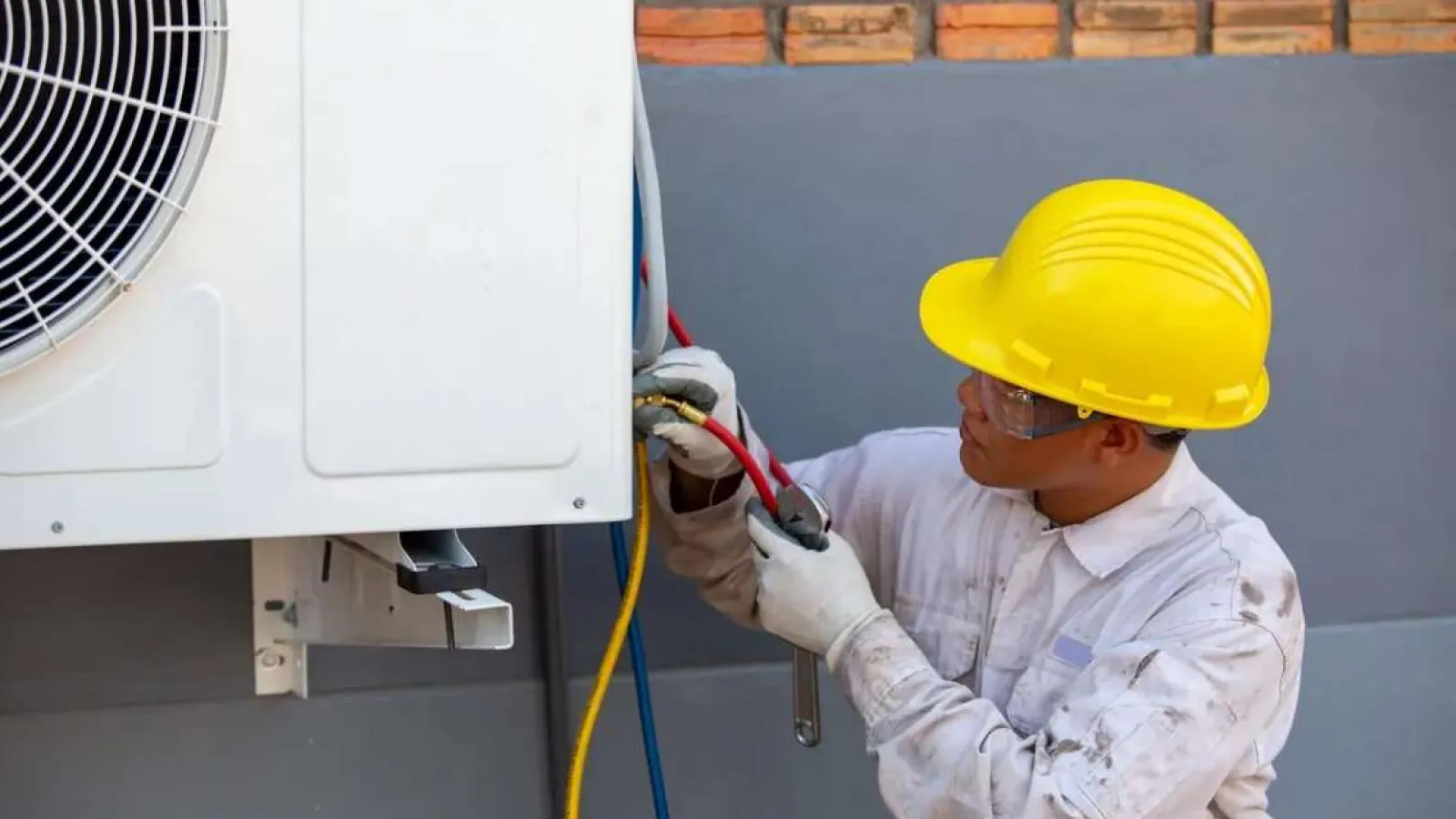 Technician wearing yellow hard hat and safety glasses servicing an outdoor air conditioning unit with tools.