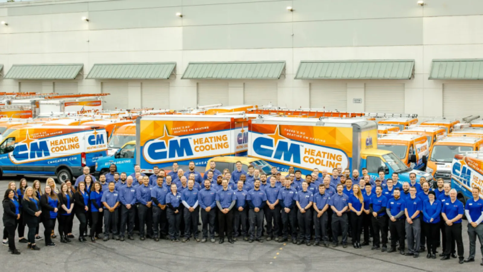 Large group of CM Heating Cooling employees posing in front of branded service trucks outside a warehouse