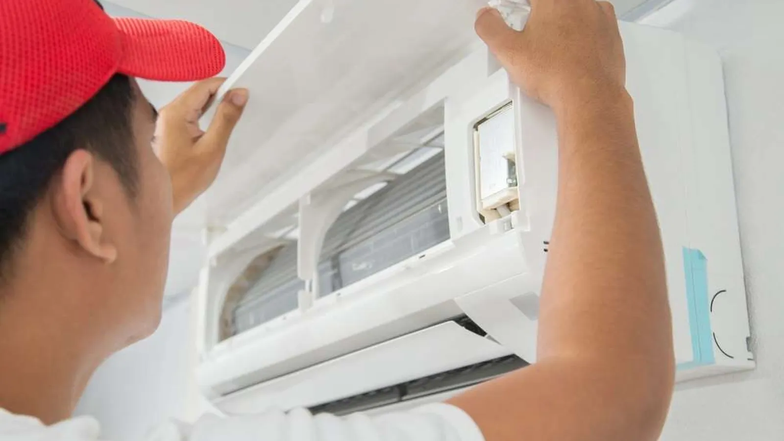 Technician wearing red cap installing or inspecting a wall-mounted air conditioning unit indoors.