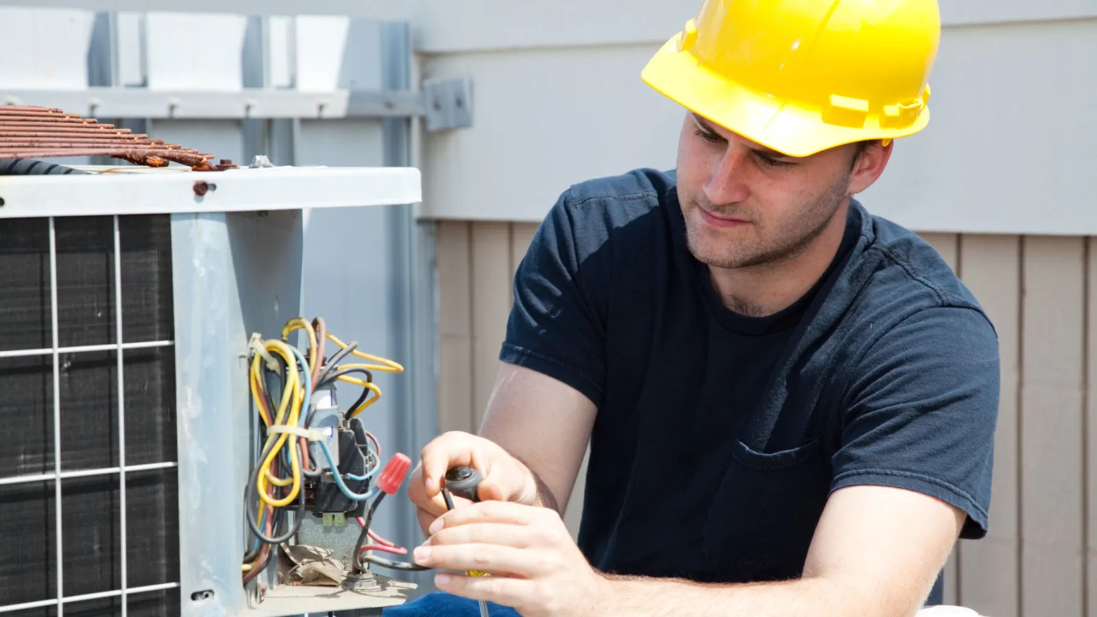 Technician wearing yellow hard hat repairs outdoor air conditioning unit with screwdriver and wires.