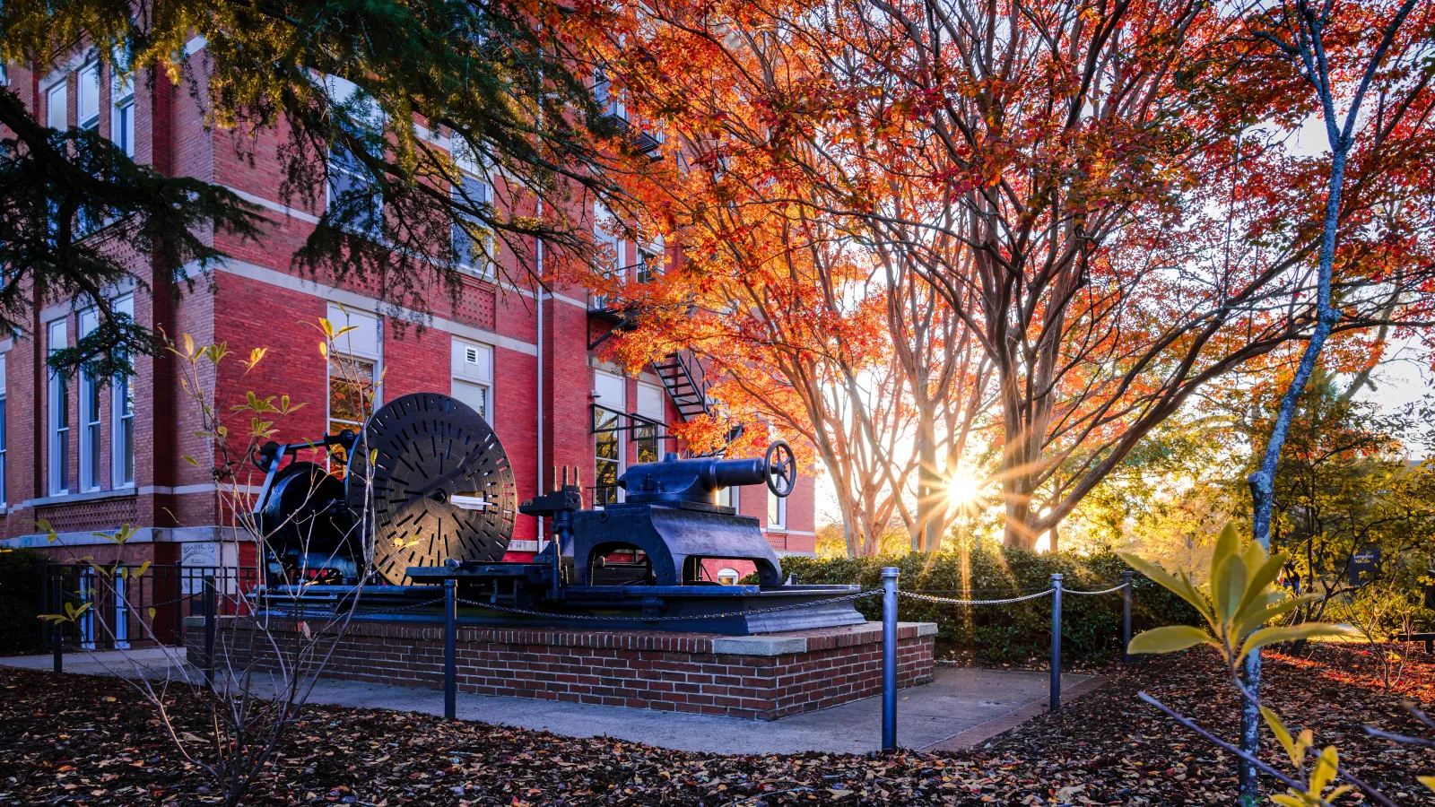 Historic industrial machinery displayed outdoors near red brick building under vibrant autumn trees at sunset