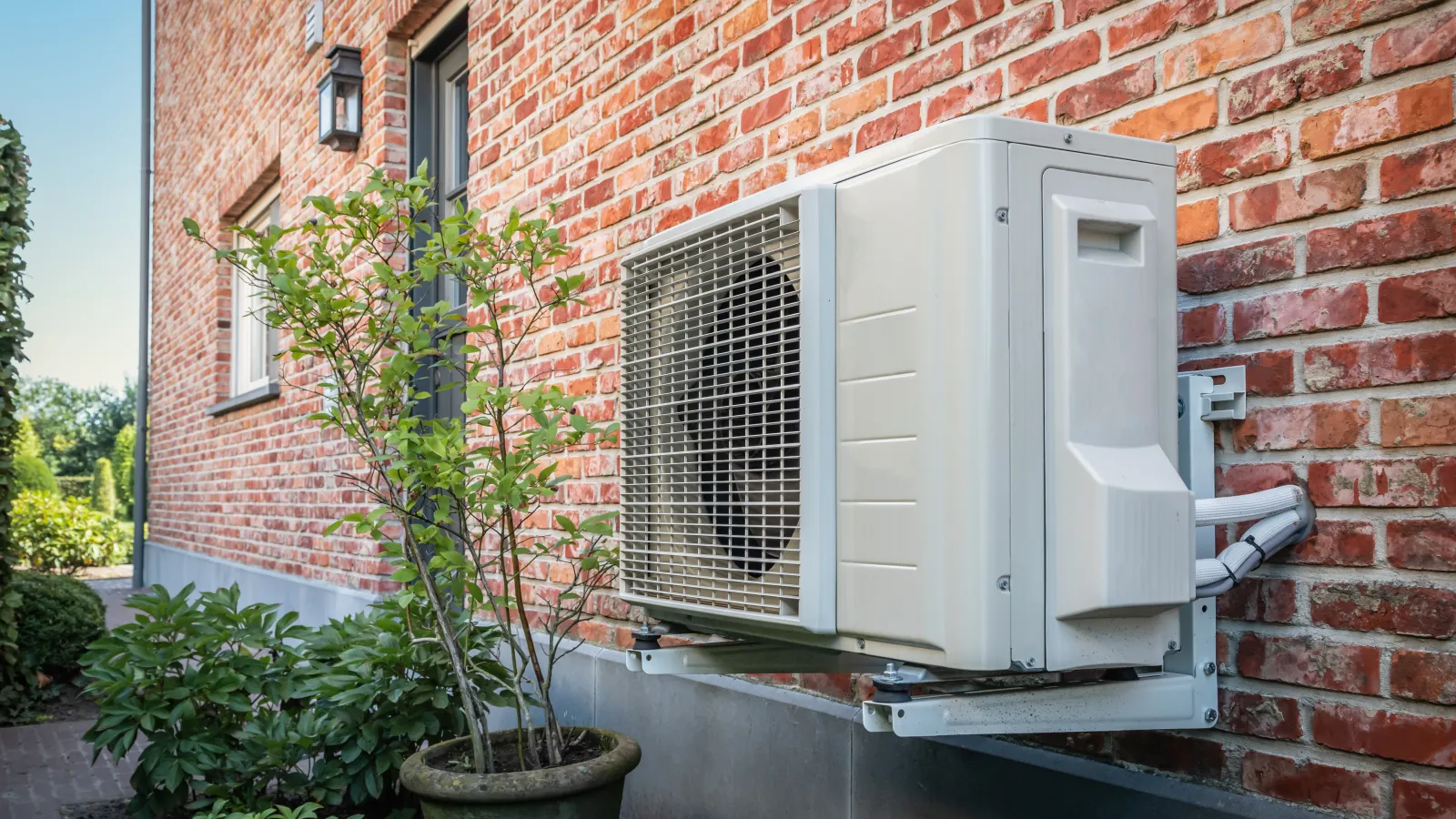 Outdoor air conditioning unit mounted on brick wall next to green potted plants in a residential garden.