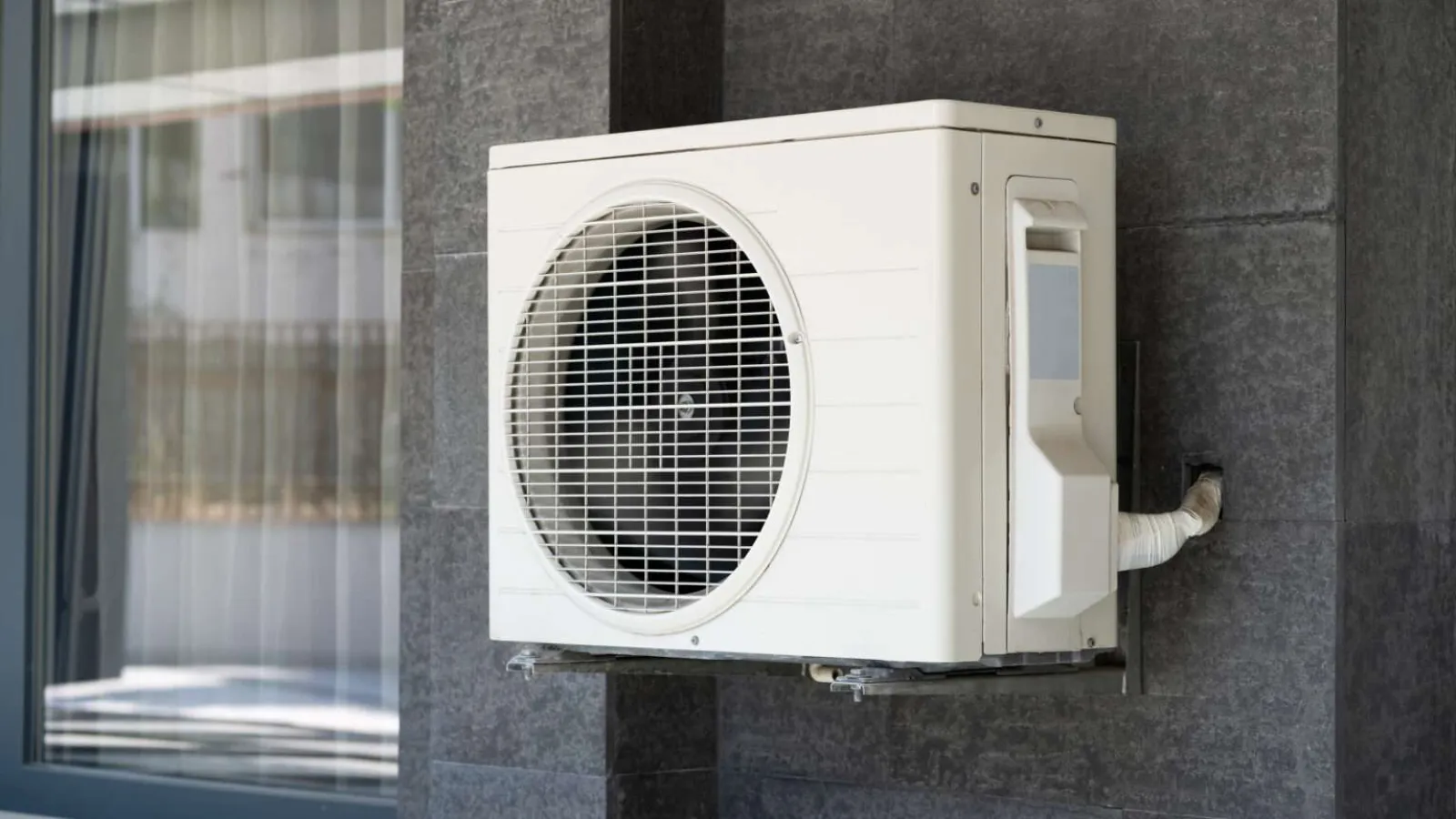 White air conditioning unit mounted on modern gray tiled exterior wall beside glass window.