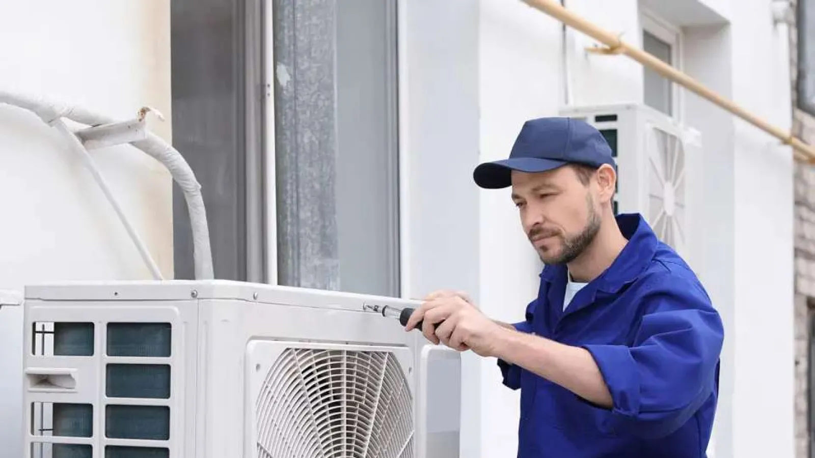 Technician in blue uniform repairing an outdoor air conditioning unit with a screwdriver beside a white building.