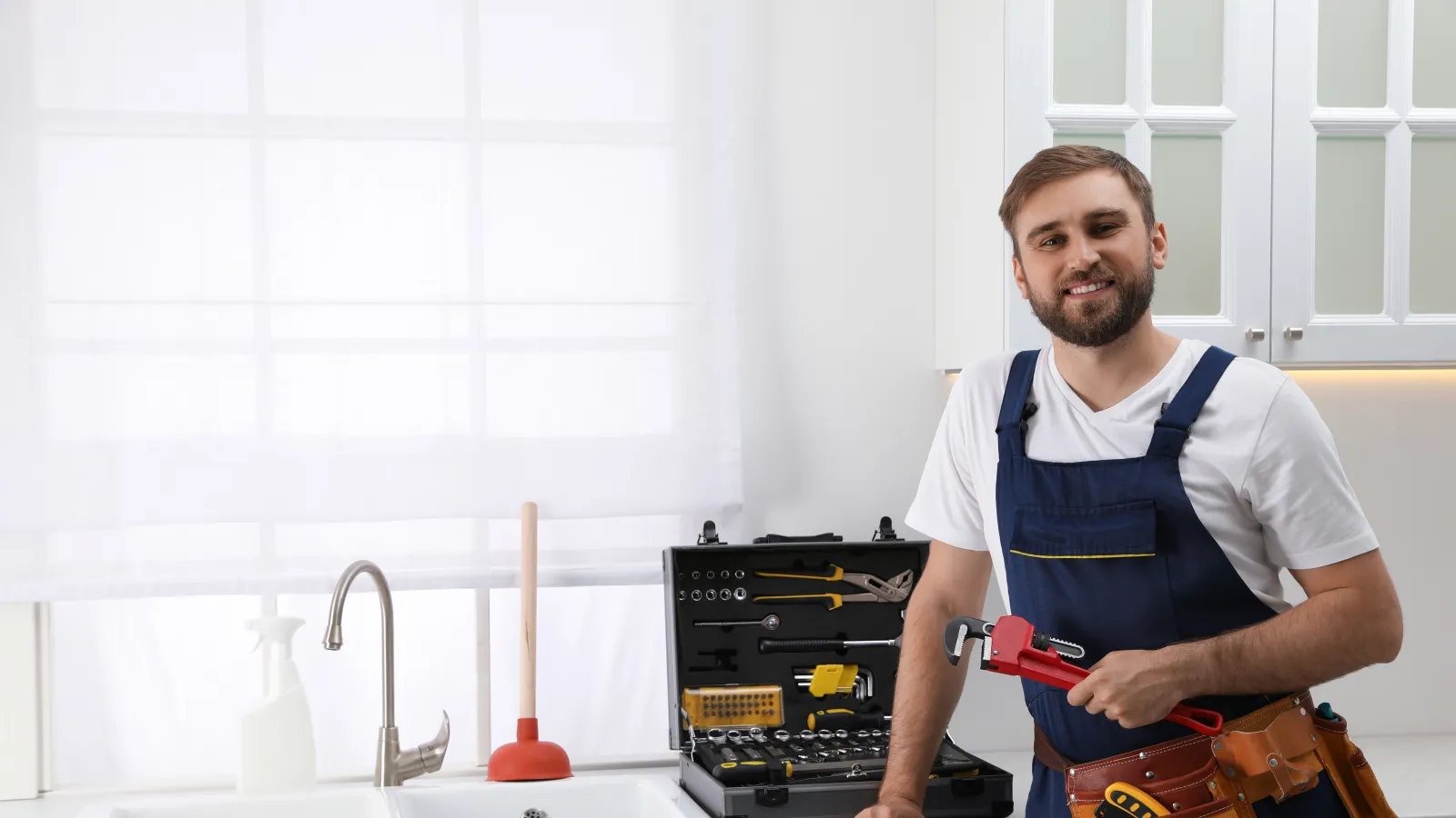 Smiling plumber in overalls holding a pipe wrench next to a sink with tools and cleaning gloves in a bright kitchen.