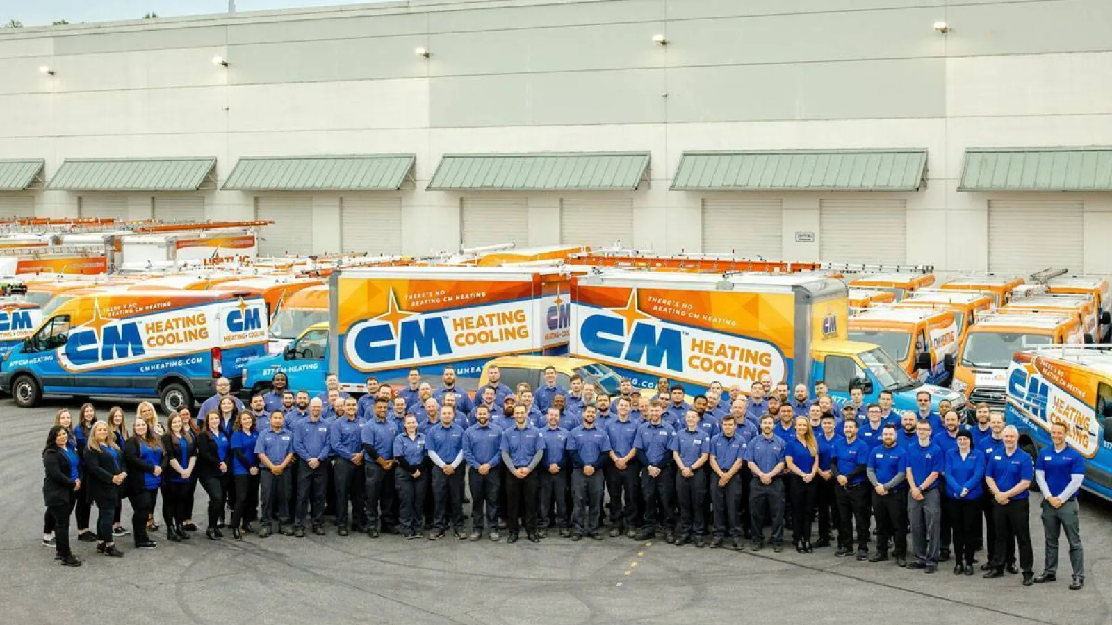Group photo of CM Heating and Cooling staff with branded service trucks in front of a warehouse building.