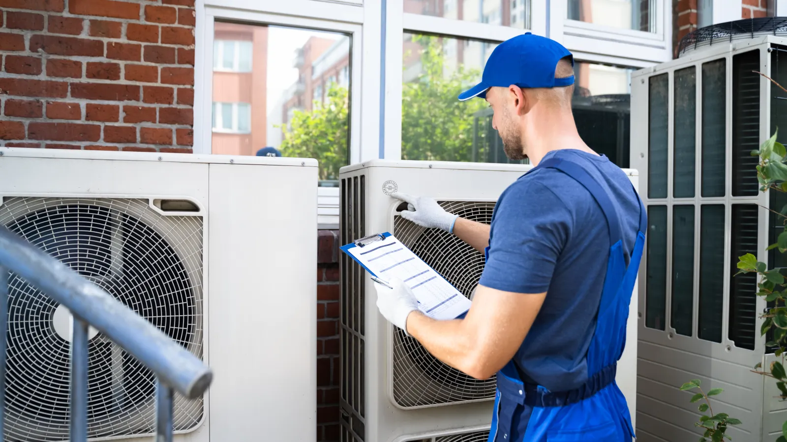 Technician in blue uniform inspecting outdoor air conditioning units with clipboard near brick wall and windows.
