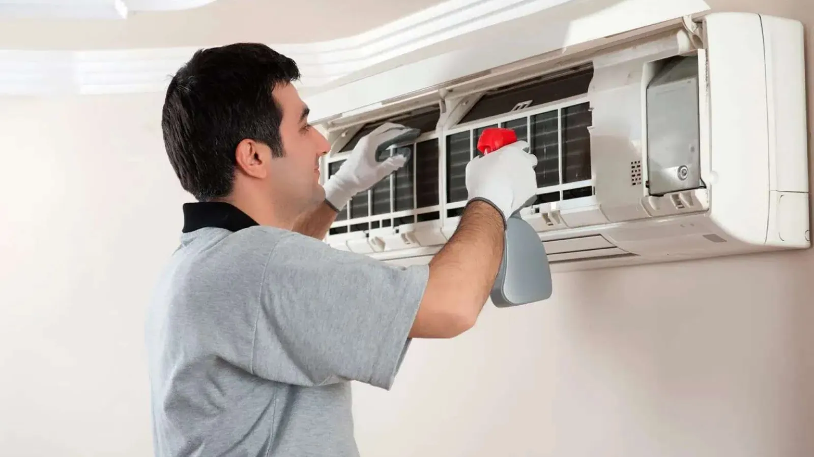 Technician cleaning and maintaining a wall-mounted air conditioning unit indoors with spray bottle and gloves.