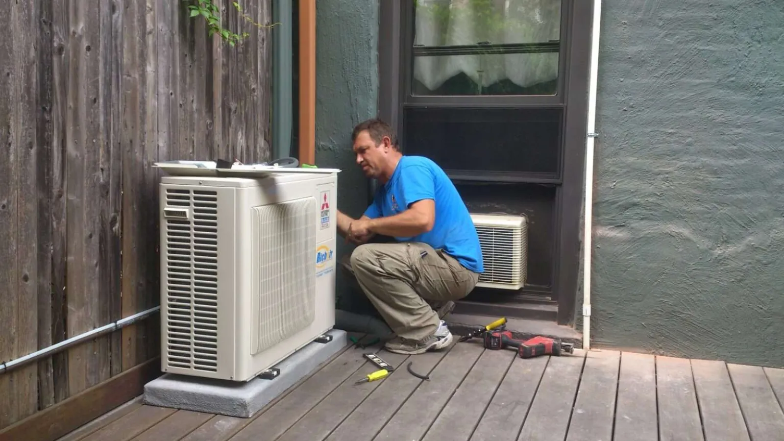 Technician with glasses inspecting and repairing an outdoor HVAC unit during daytime.