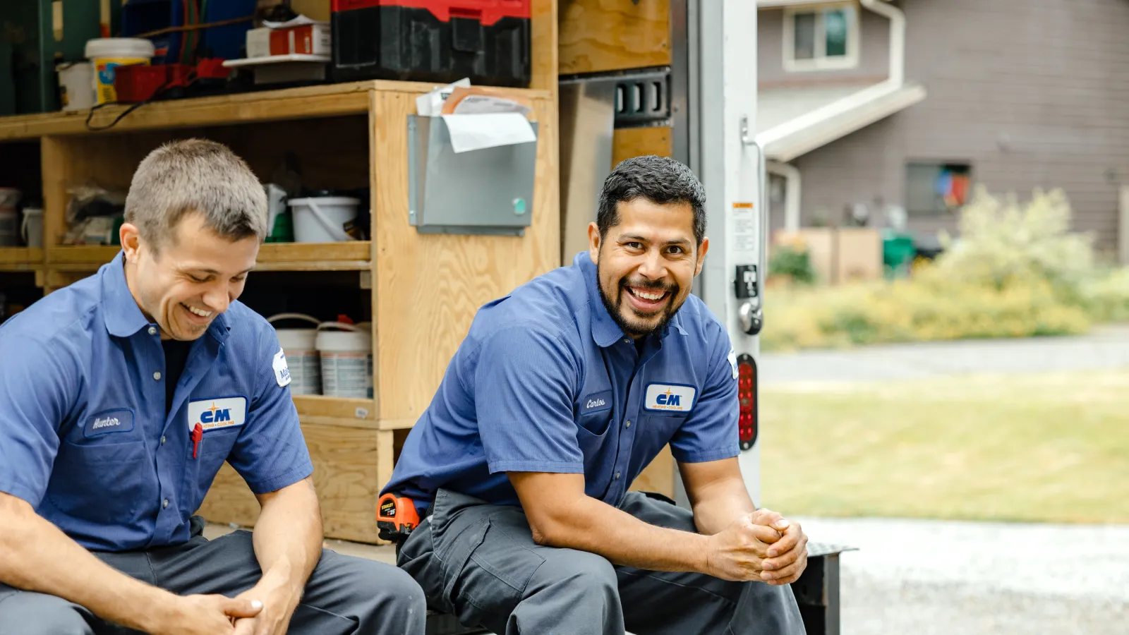 Two smiling technicians in blue uniforms sitting on the back of a work van with tools around them.