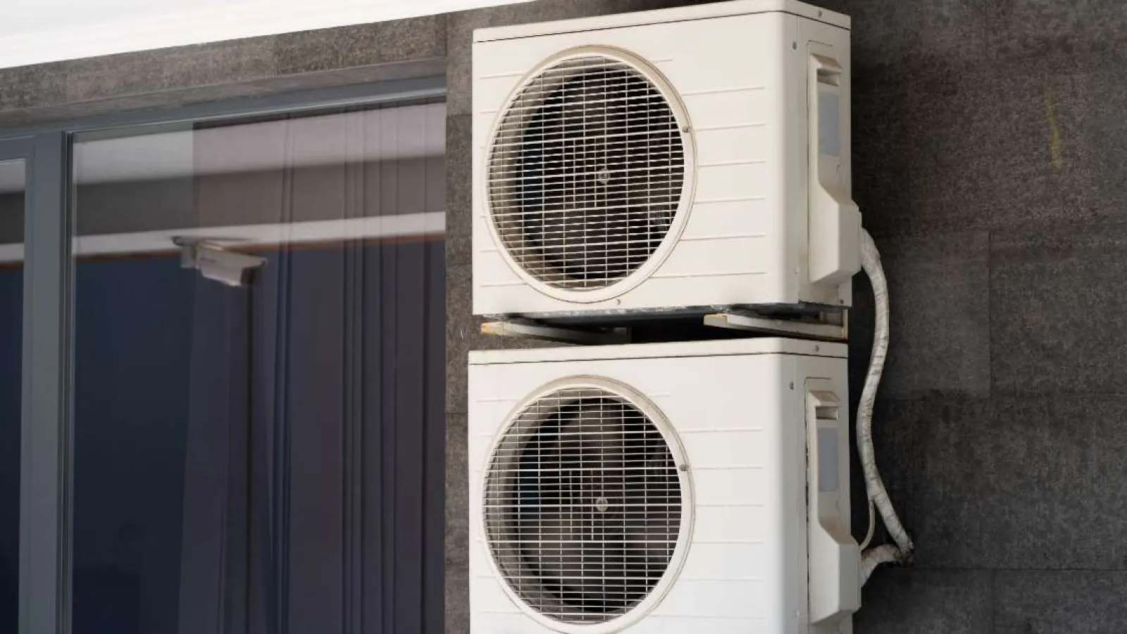 Two white air conditioning units mounted vertically on an exterior dark stone wall beside a glass window.