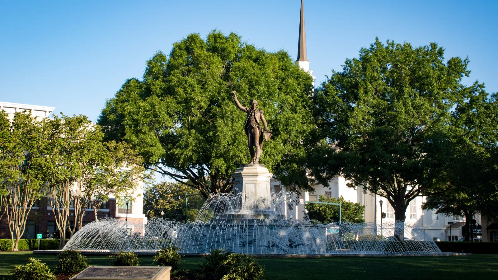 Historic statue with raised arm surrounded by a large fountain and lush green trees under clear blue sky.