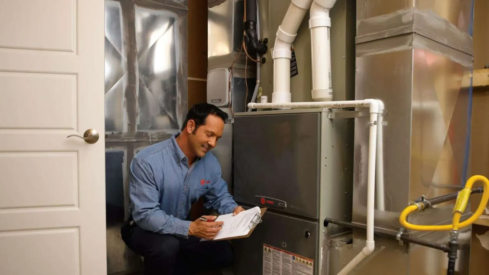 Technician inspecting and taking notes on a residential HVAC furnace system in a utility room.