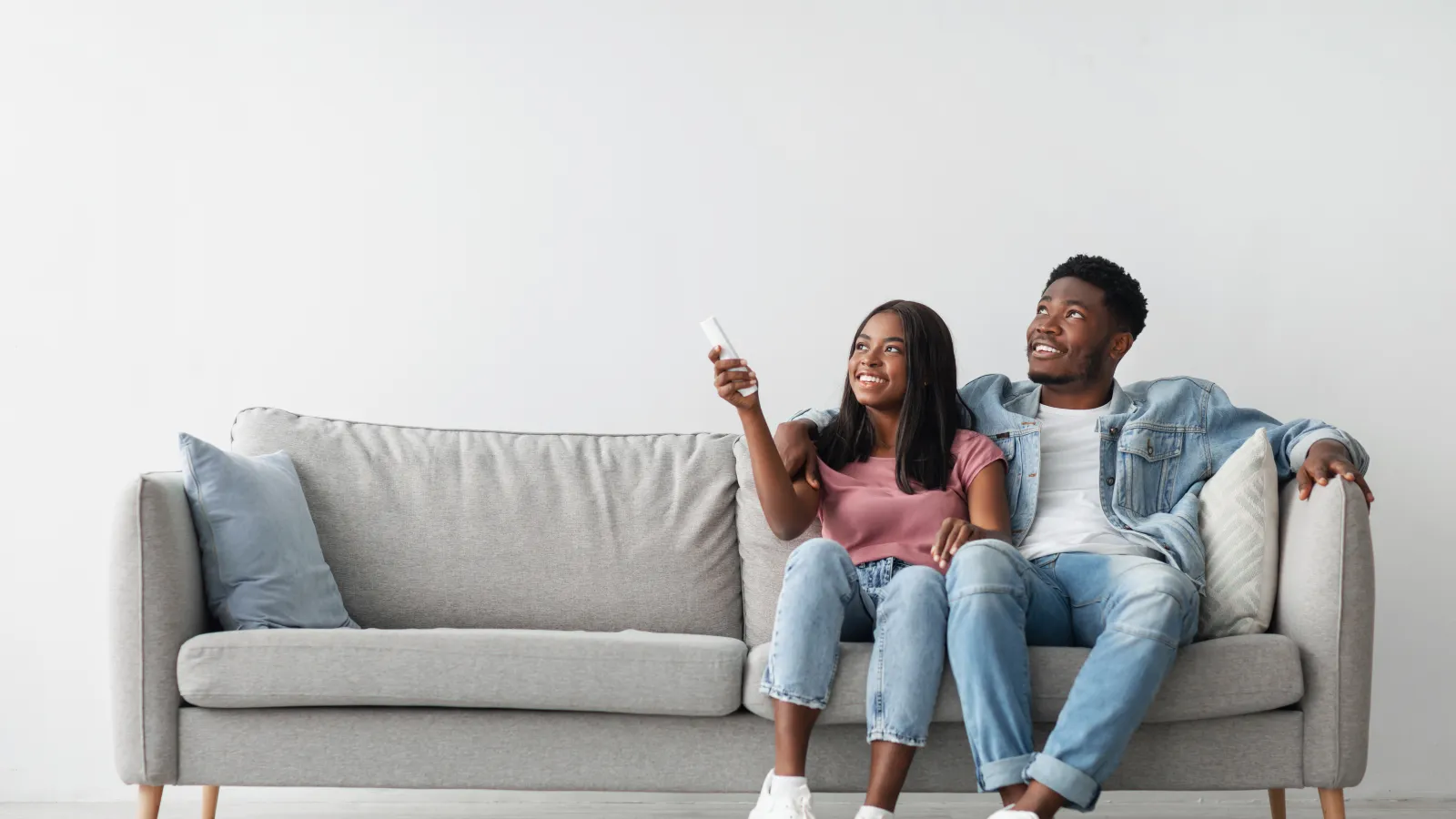 Happy young couple sitting on a gray couch watching TV with remote control in a bright modern living room.