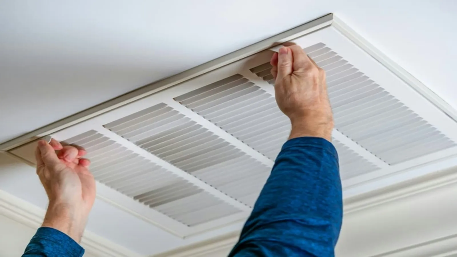 Person wearing blue long sleeves adjusting a white ceiling air vent grille in a room.