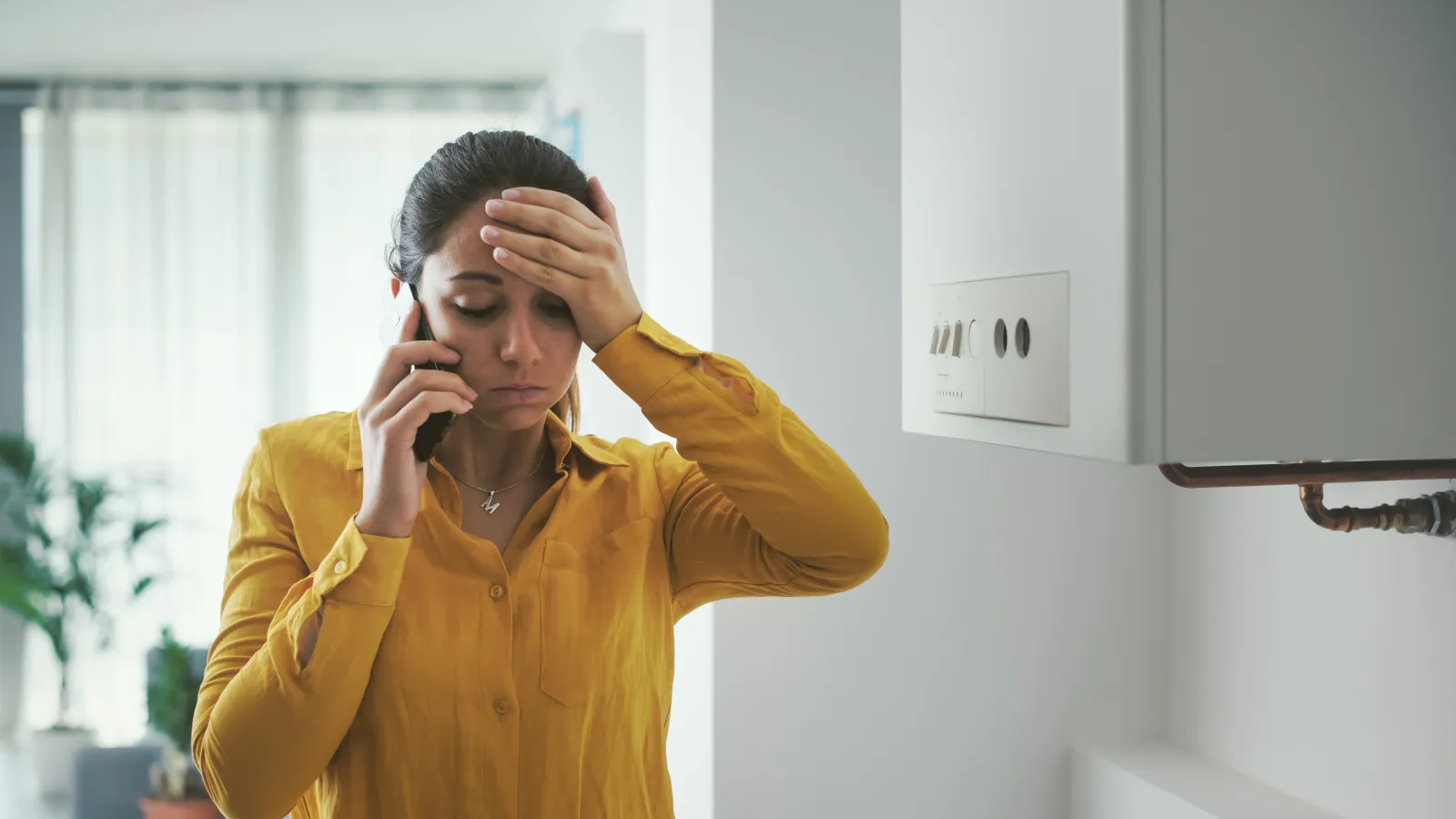 Worried woman in yellow shirt on phone next to a boiler, holding her head in frustration inside a bright room.