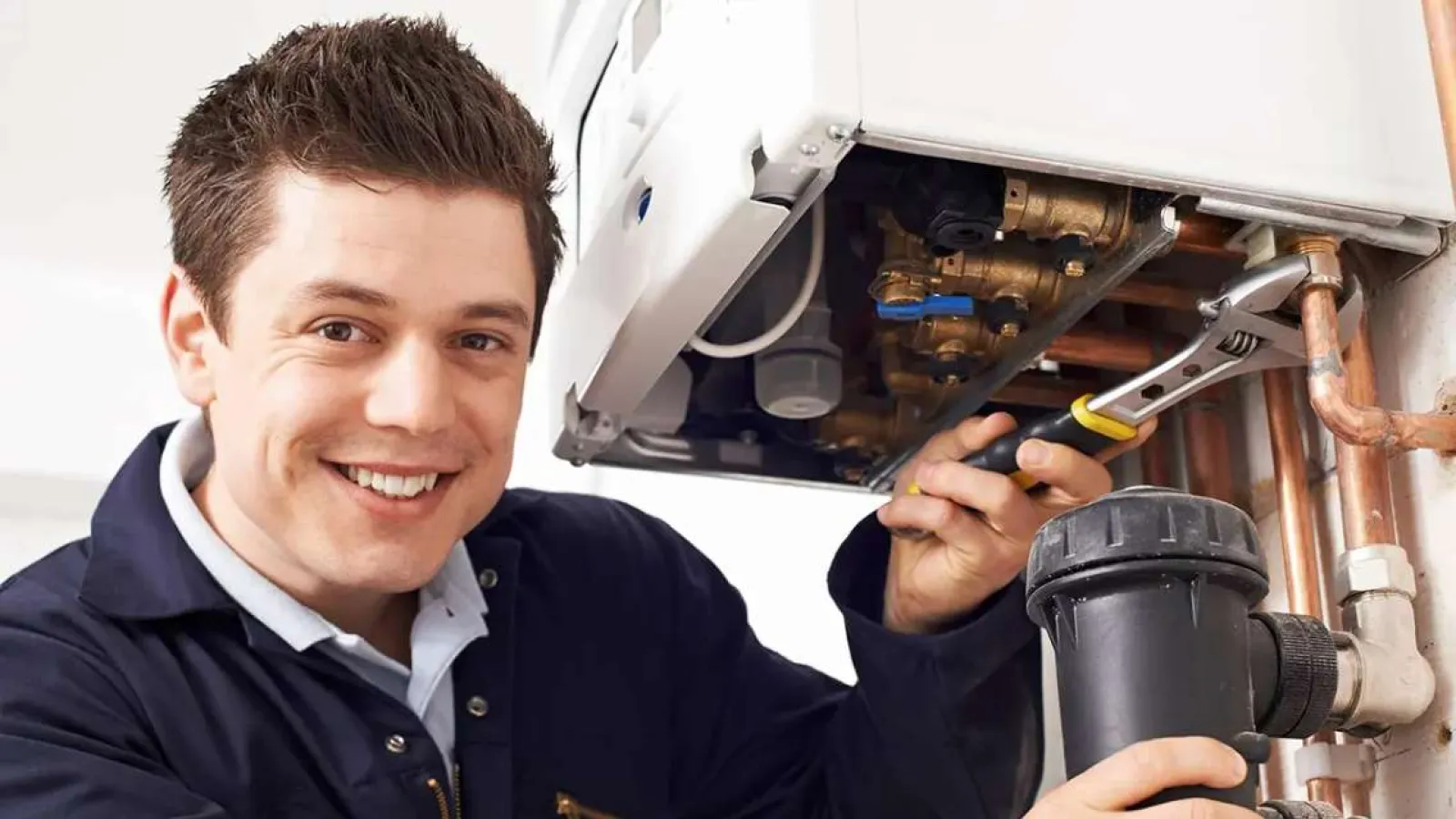 Smiling plumber fixing boiler pipes with a wrench in a residential setting wearing navy work uniform