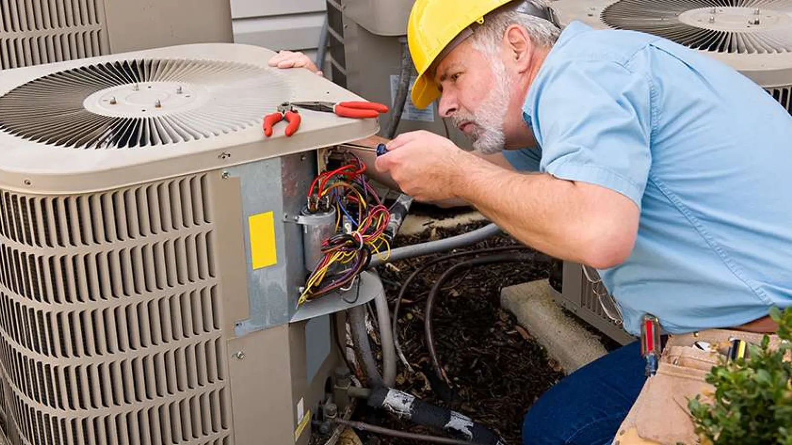 Technician wearing a yellow hard hat repairing an outdoor air conditioning unit with exposed wiring.