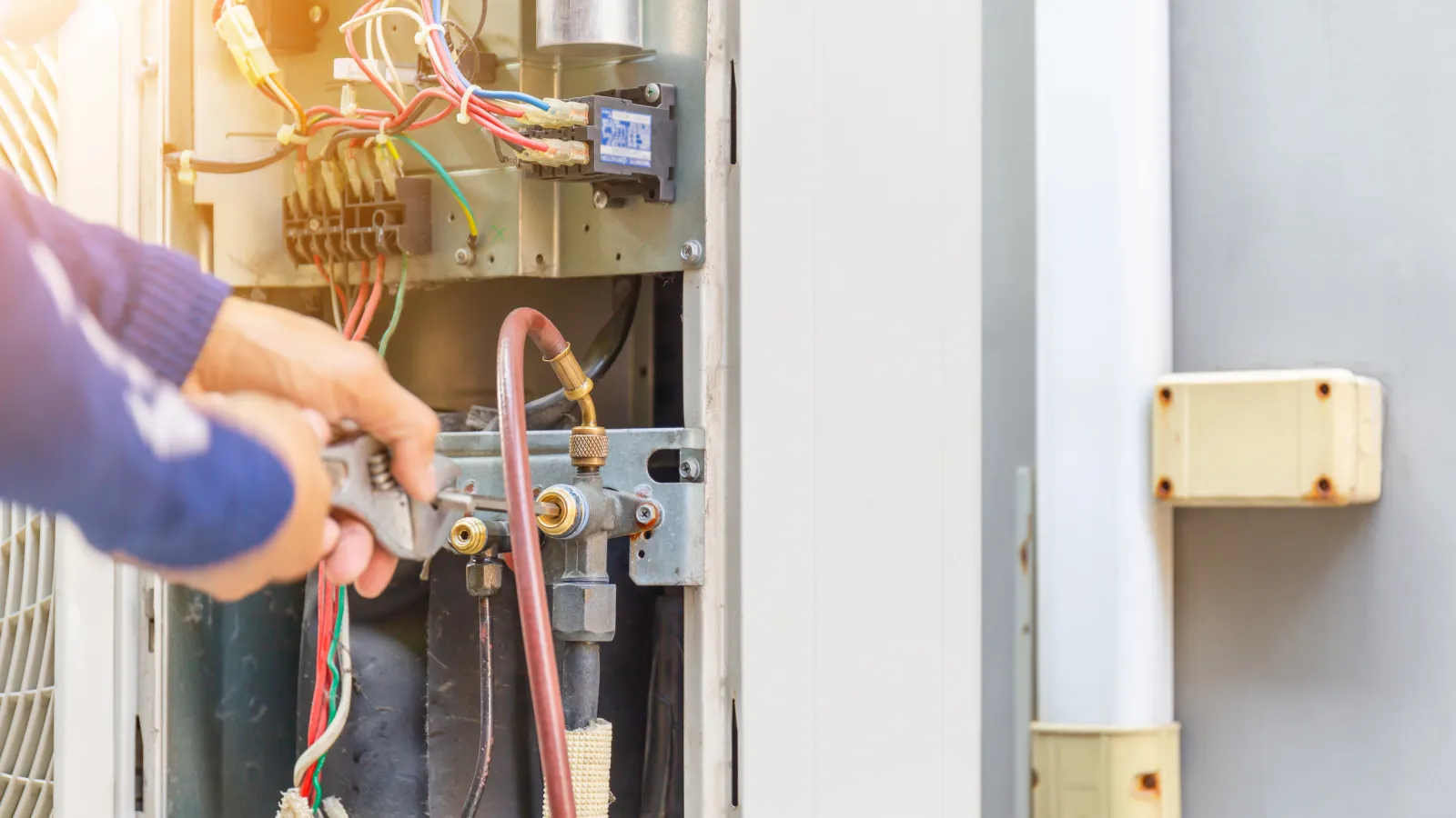 Technician repairing and maintaining an air conditioning unit with tools and wiring.