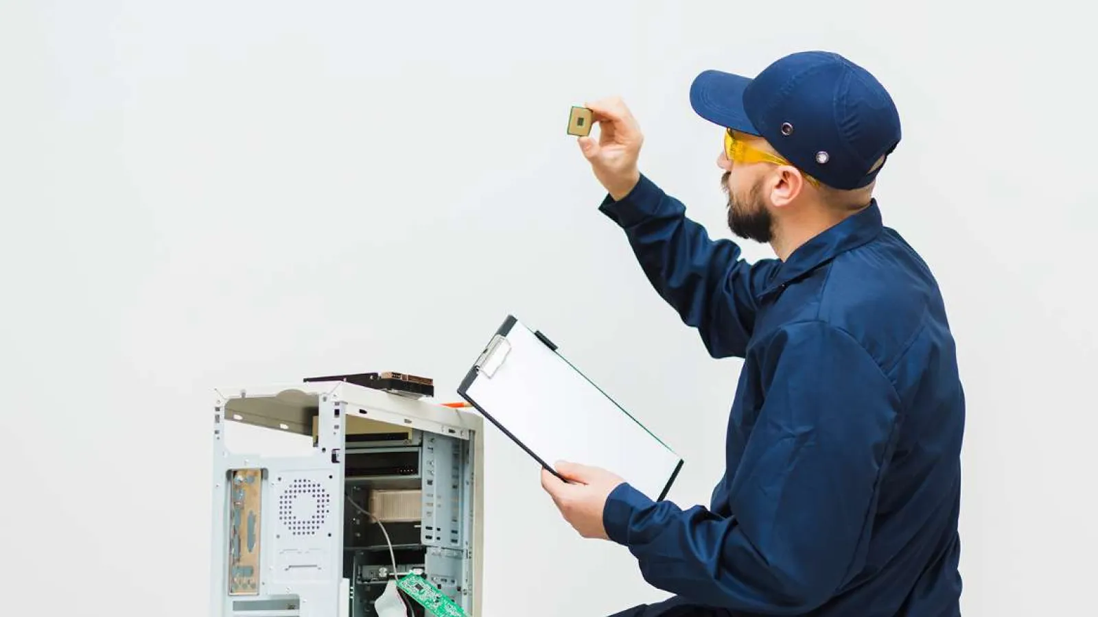Technician in navy uniform holding a CPU and clipboard beside an open desktop computer tower in a white room.