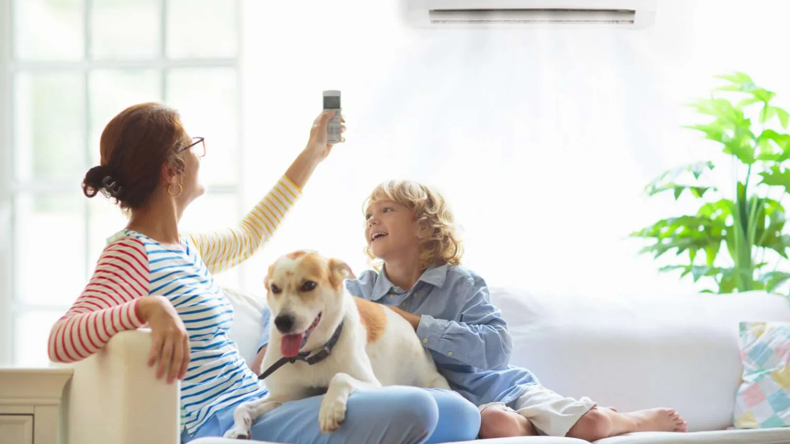 Mother using remote to control air conditioner while child and dog relax on sofa in bright living room