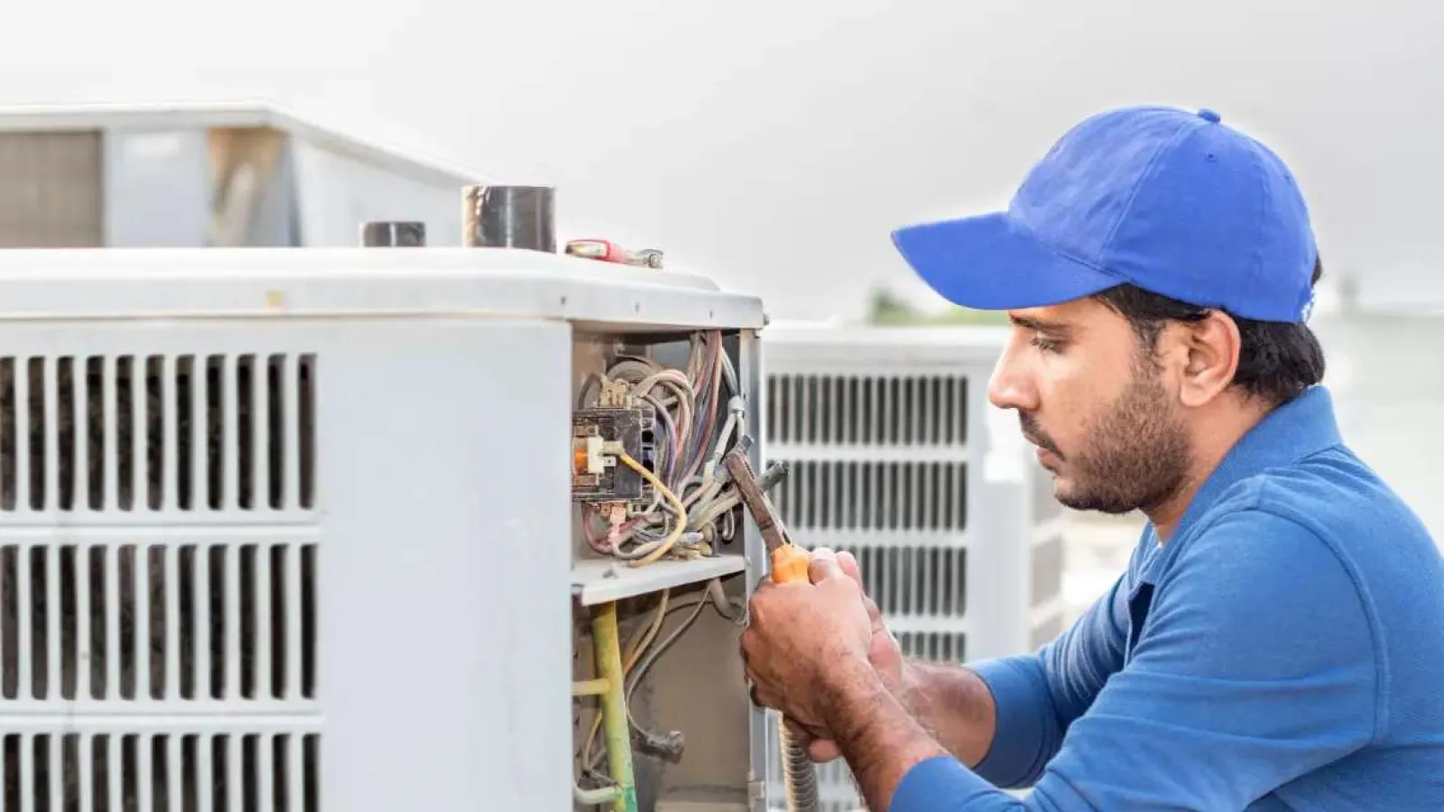 Technician in blue uniform repairing an outdoor air conditioning unit with tools on a rooftop.