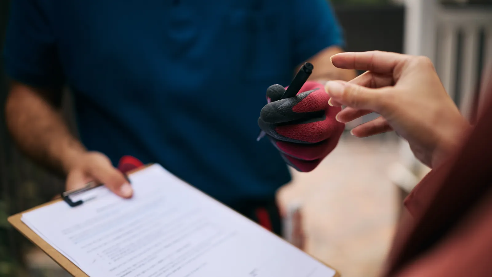 Delivery person in gloves handing a pen to a customer for signing a document on a clipboard outdoors.