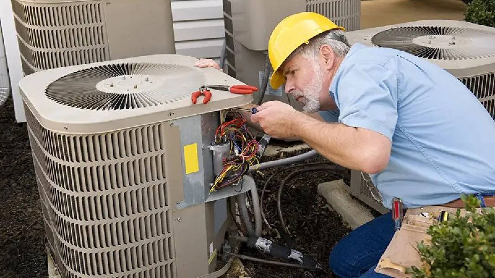 Technician in yellow hard hat repairing outdoor HVAC air conditioning unit with tools on a residential property.