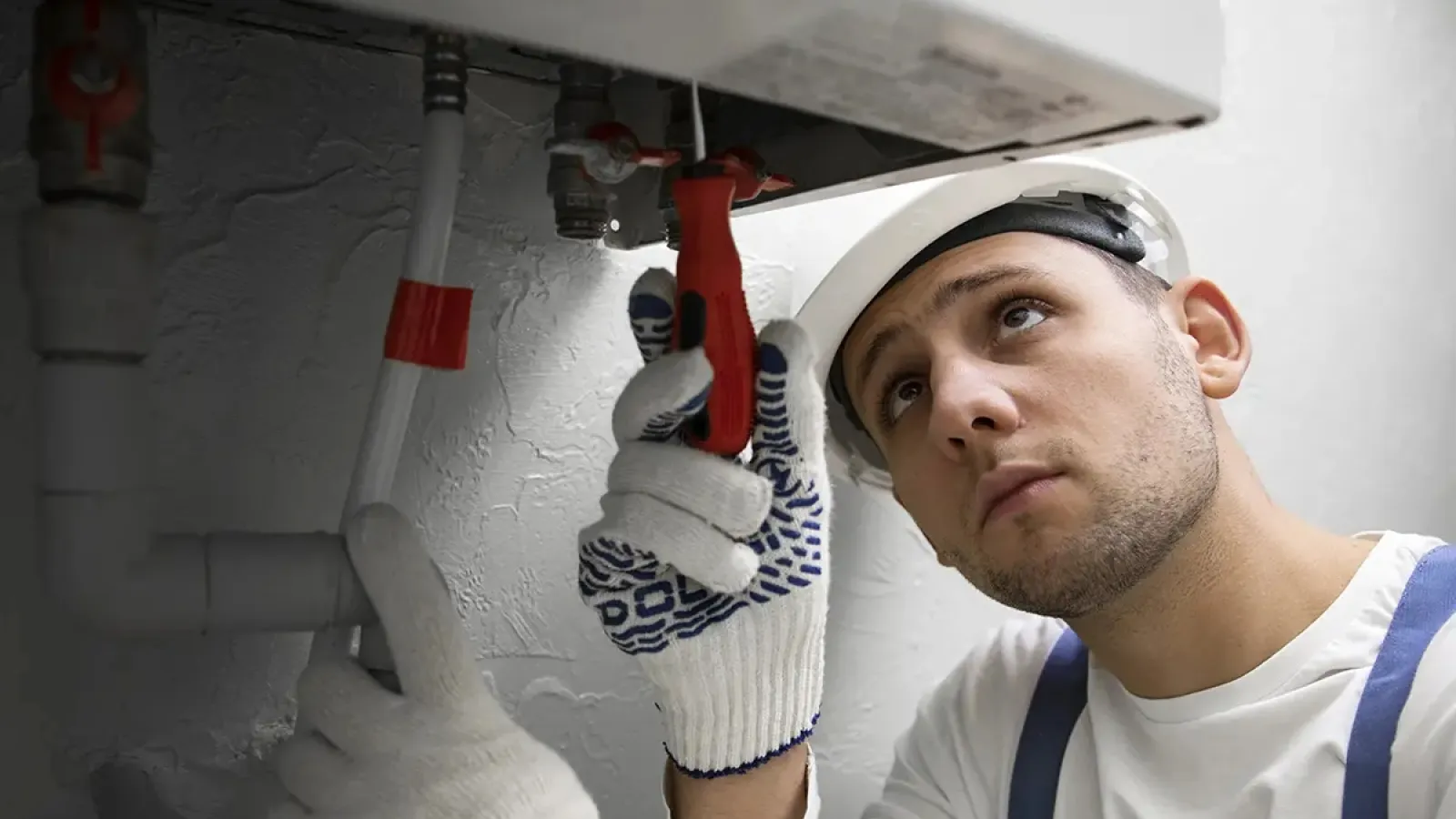 Plumber in white helmet and gloves adjusting a valve under a sink with white pipes in a utility room.