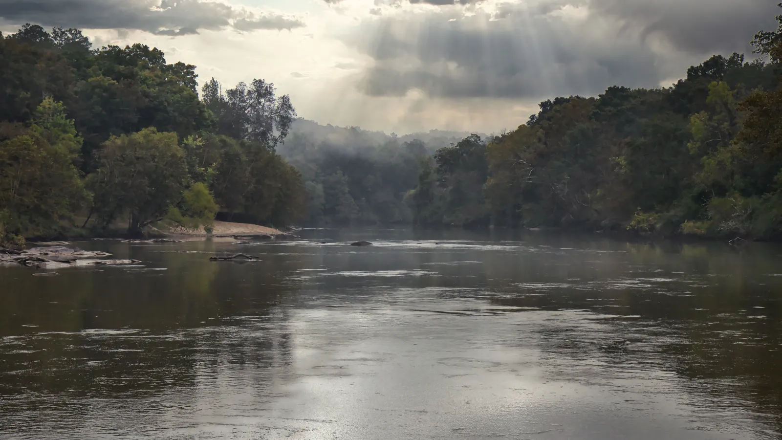 River flowing through dense forest under cloudy sky with sun rays breaking through the clouds.