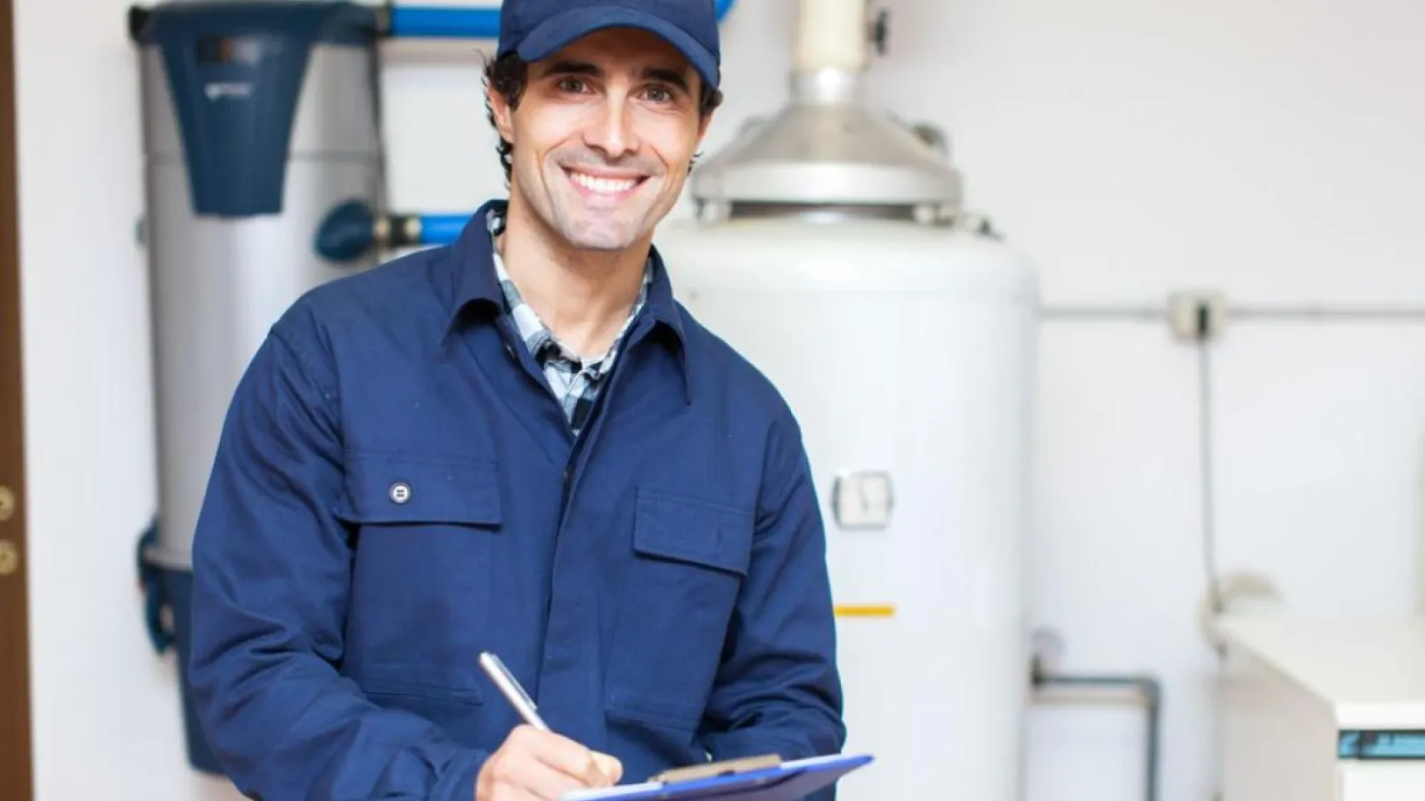 Smiling technician in navy uniform holding clipboard standing near water heater and pipes indoors.