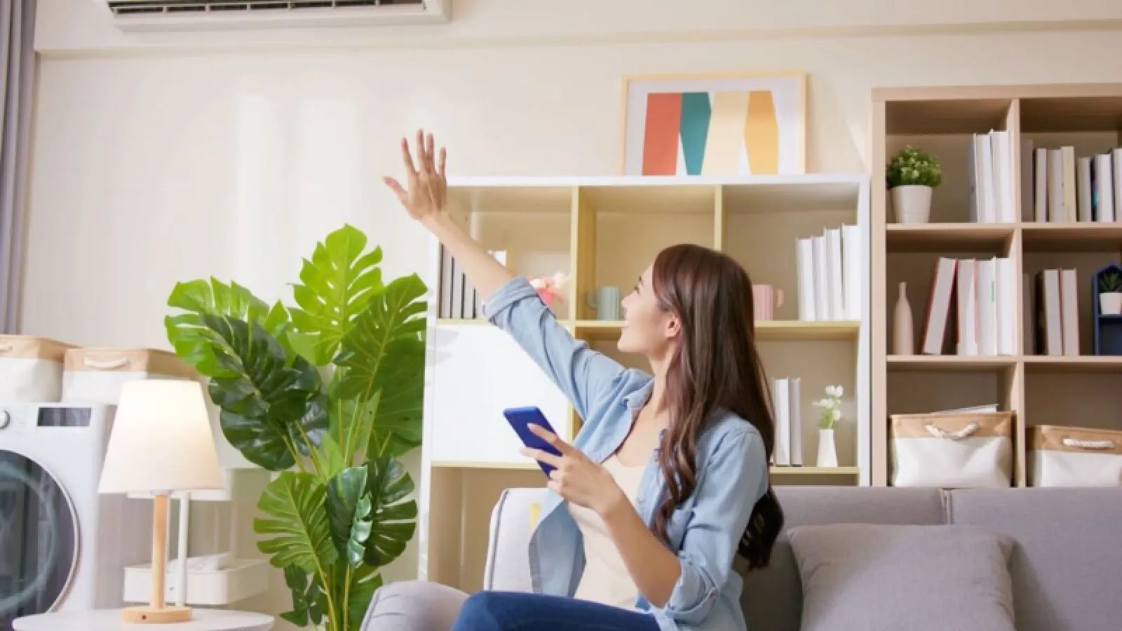 Woman holding smartphone setting remote control for wall-mounted air conditioner in cozy living room.