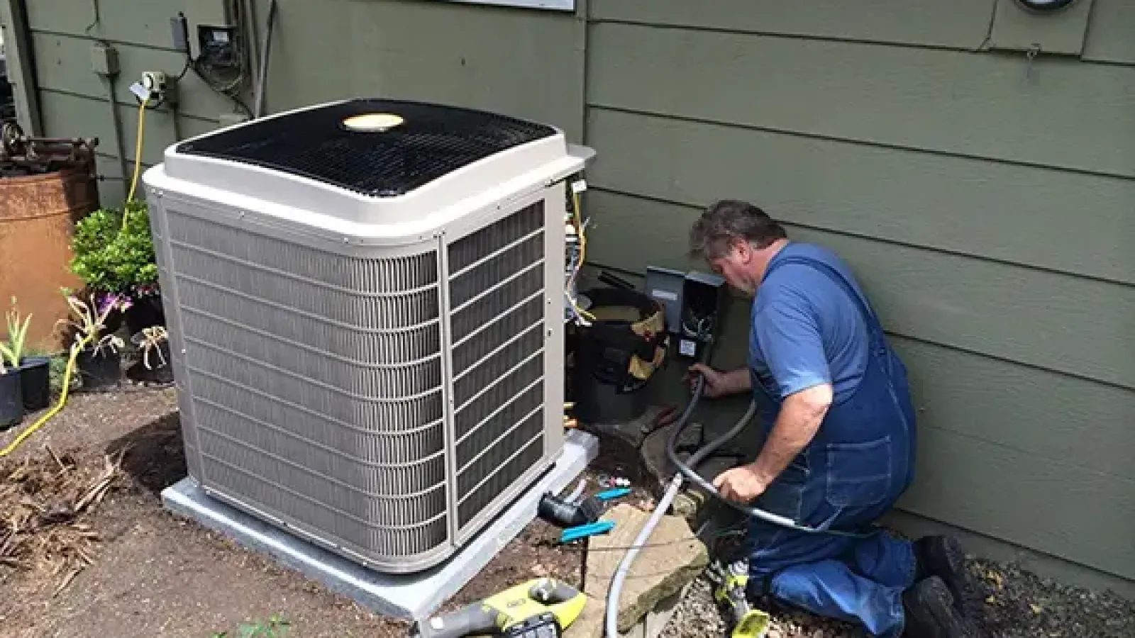 Technician in blue overalls repairing a residential air conditioning unit outside a green house on a gravel surface.