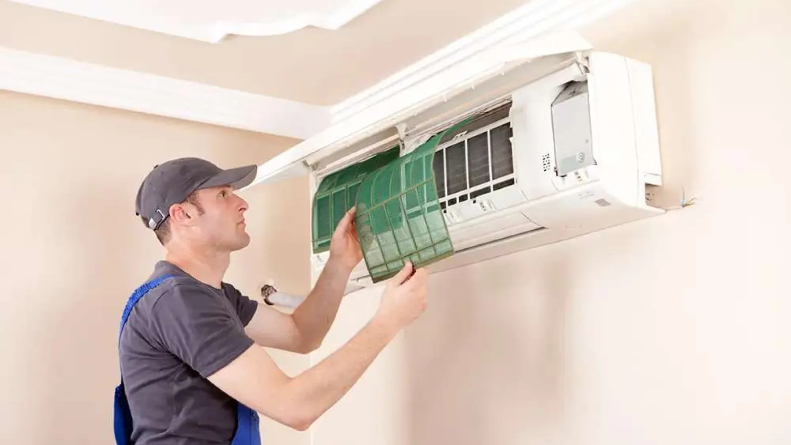 Technician inspecting and cleaning air conditioner filter inside a wall-mounted unit in a beige room.